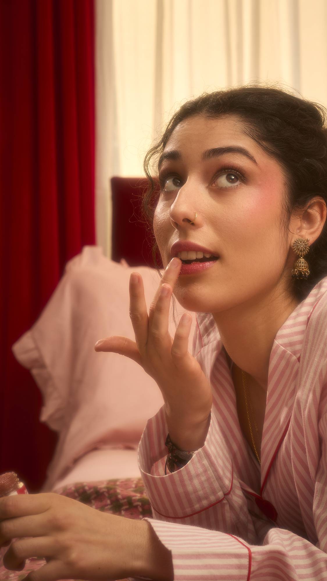 A woman in pink striped pyjamas looks upward as she applies a shimmering Cherry Bubblegum lip scrub to her bottom lip with her finger. She is wearing ornate gold earrings and a nose ring while reclining in a warm, romantic bedroom setting.