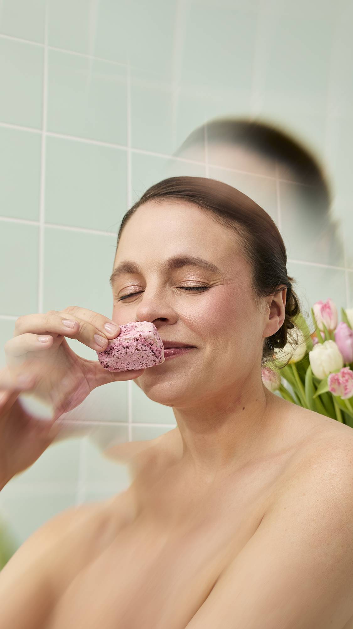 A smiling woman with dark hair holds the pink, textured "Cherry Pie" scrub bar to her nose, closing her eyes to take in the scent. The scene is set against a mint green tiled wall with blurred pink and white tulips in the background.