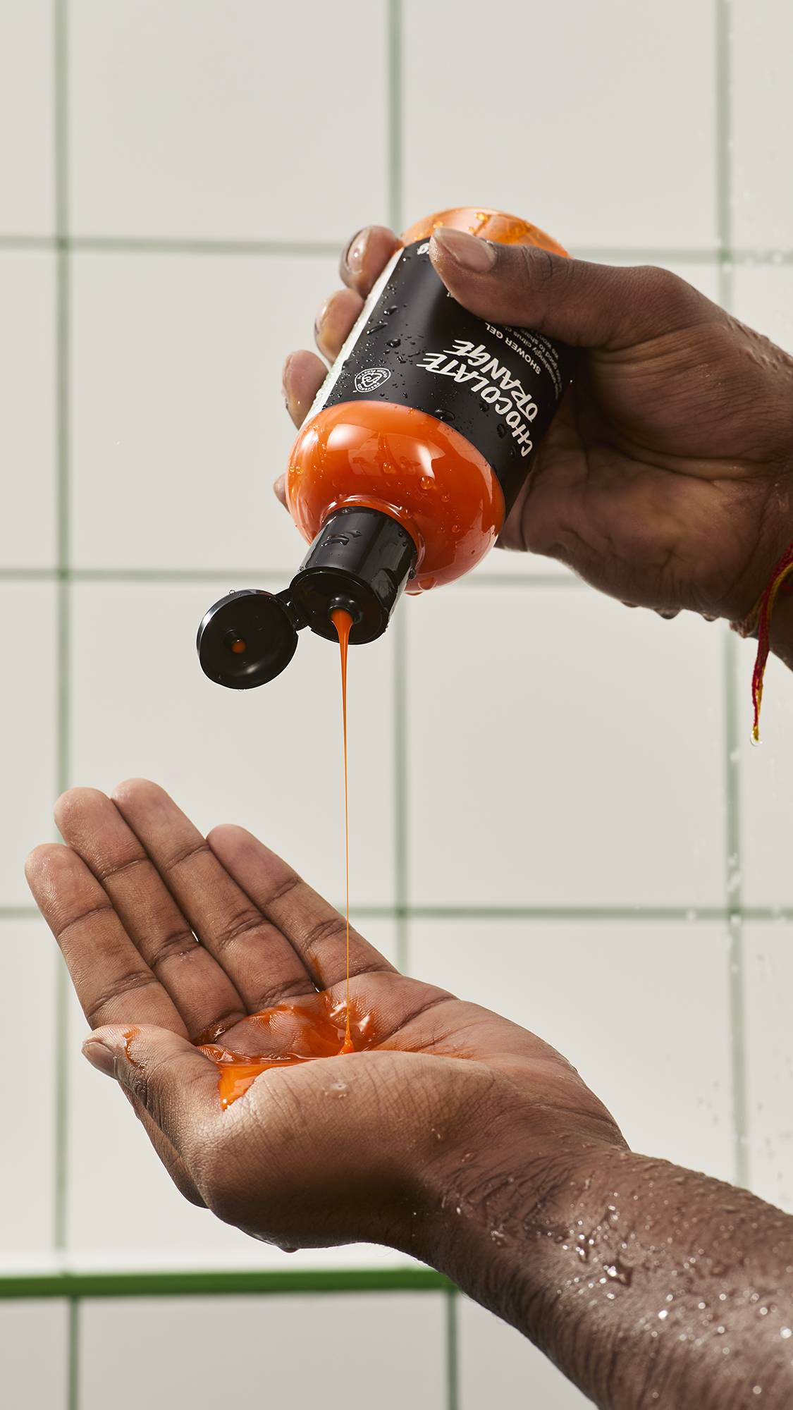 A person squeezes a bottle of Lush "Chocolate Orange" shower gel upside down, pouring a steady stream of rich orange-bronze liquid into their open palm against a white tiled wall.