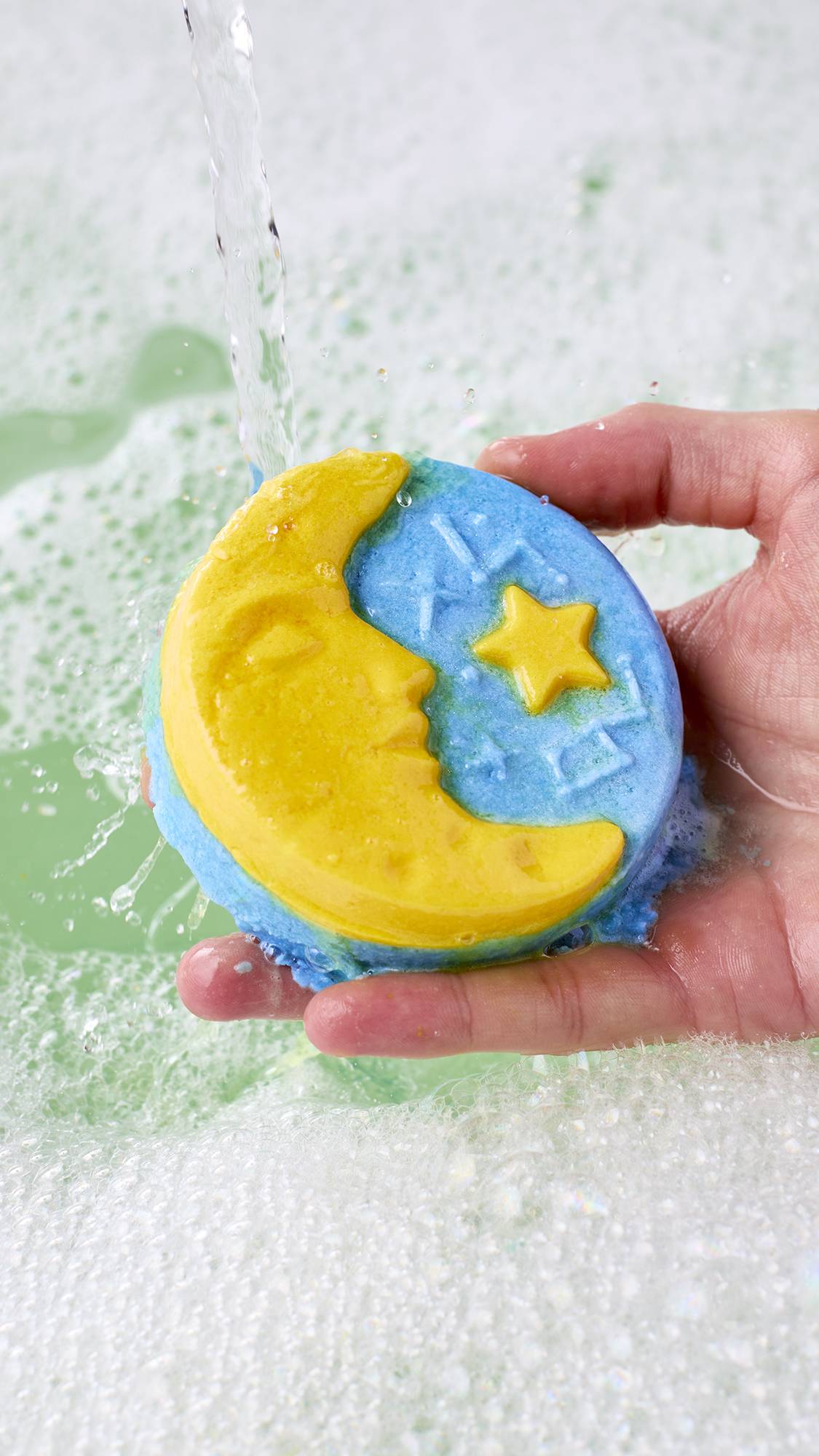 A hand holds a yellow crescent moon and blue star bubble bar under running water, fizzing gently above a surface of pale green bathwater covered in fine white bubbles.
