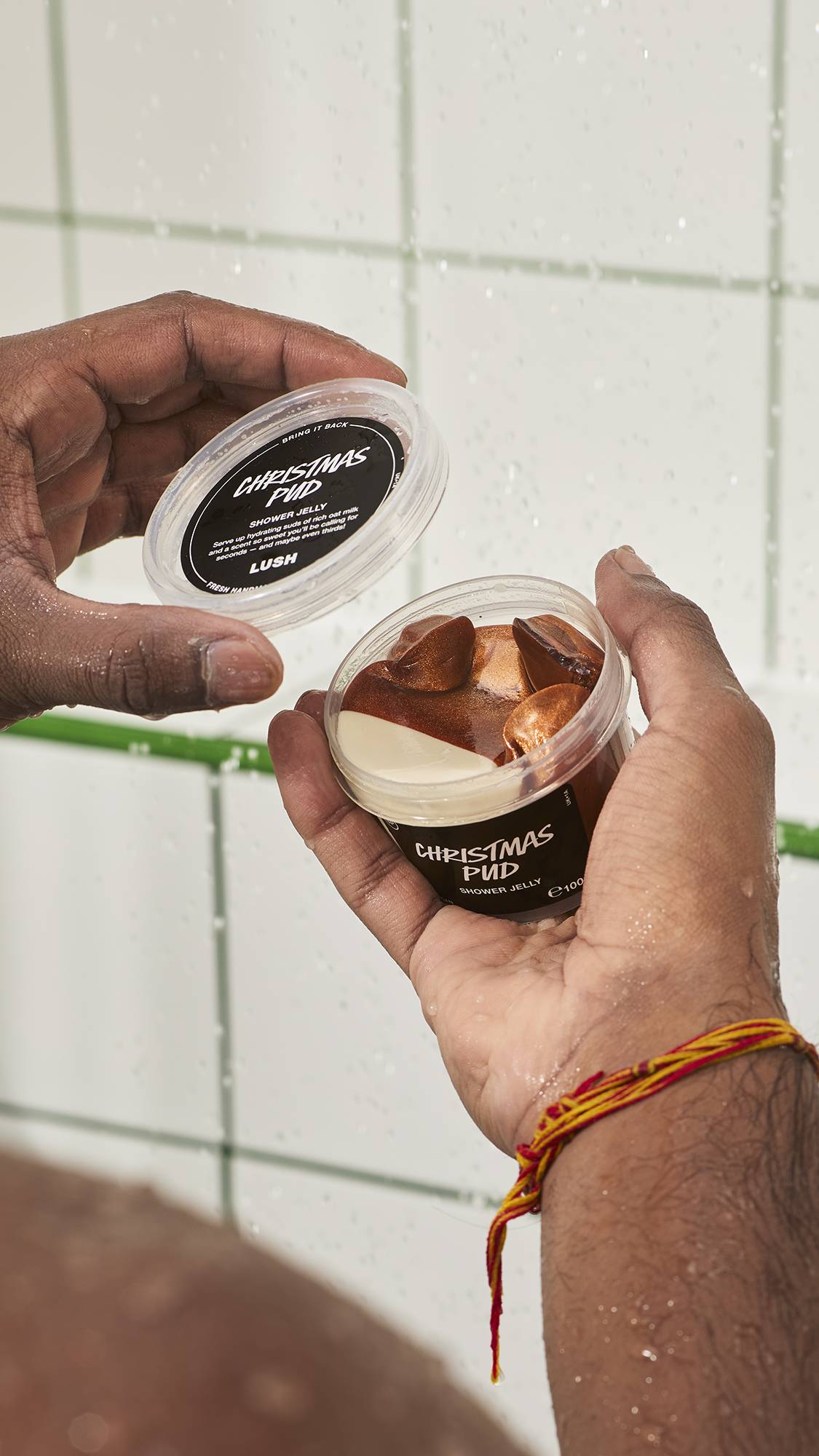 Hands holding an open plastic tub of Lush "Christmas Pud" shower jelly in the shower, revealing a bronze-gold character jelly inside against a white tiled wall background.