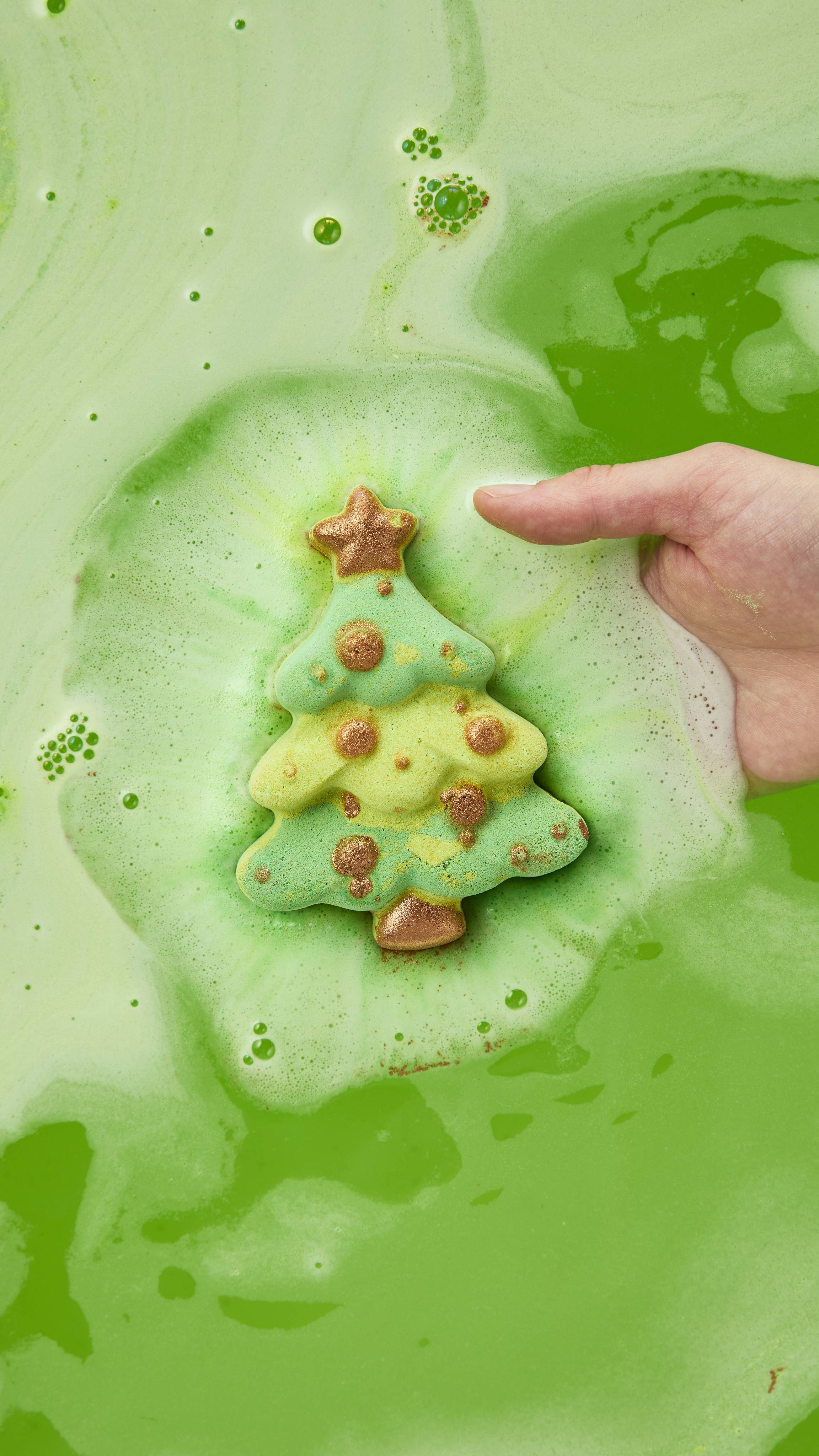 The image shows a hand gently placing a green Christmas tree bath bomb with gold accents into vivid green bathwater, dissolving into a circle of pale green foam