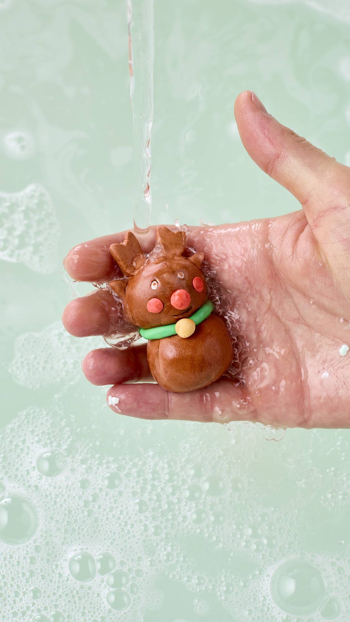 A hand holds a small brown reindeer-shaped character crafted from the Lush "Christmas Wreath" fun product, with antlers, a bright red nose, and a green collar under running tap water over pale bathwater sprinkled with white bubbles.