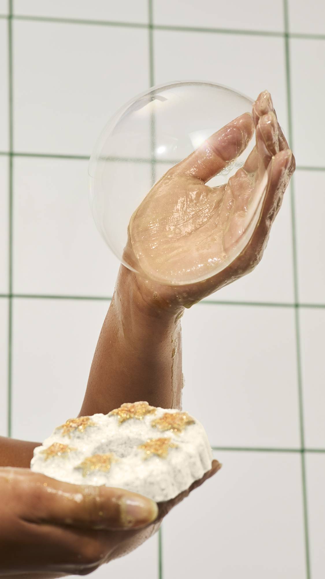 A person holds a white star-topped wreath bath product in one hand while blowing a large, clear soap bubble from their raised, wet palm against a white tiled wall.