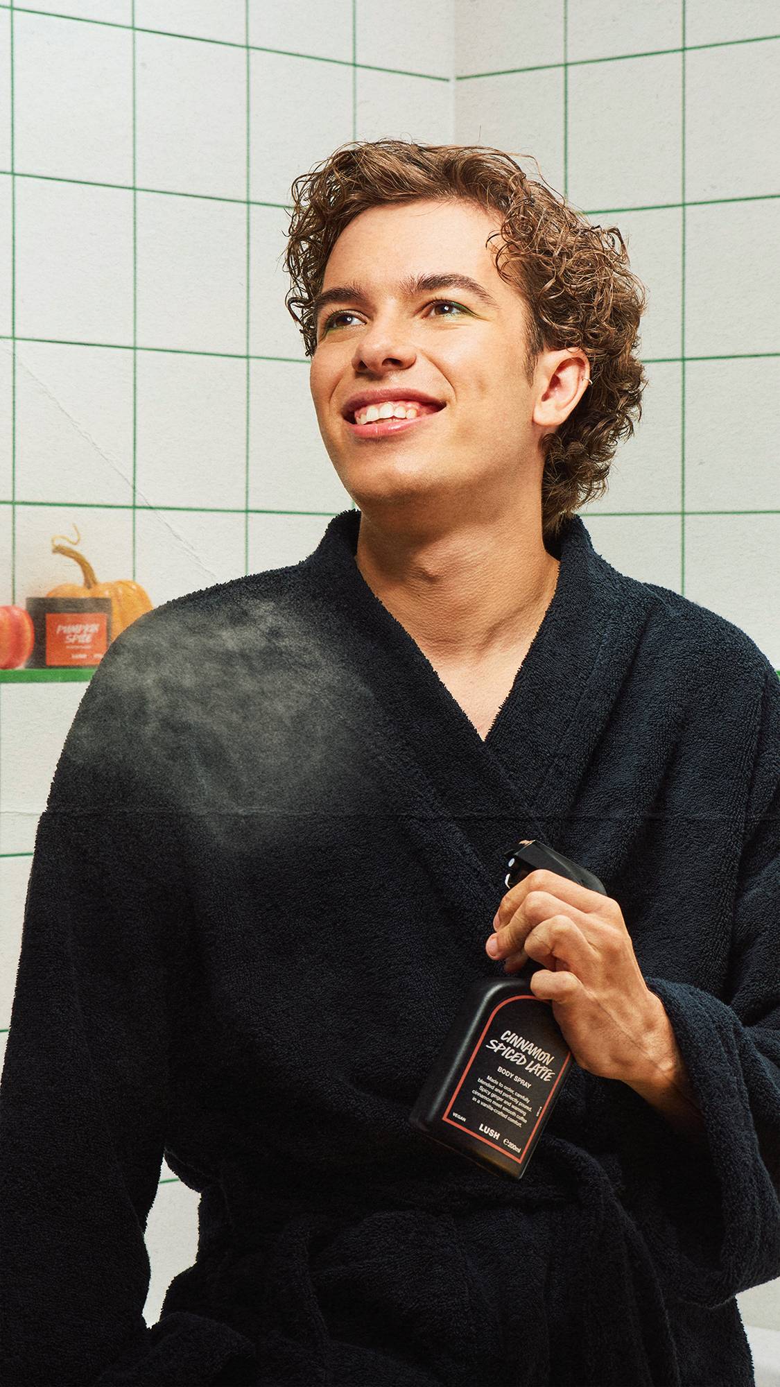 A smiling person with curly hair wearing a dark dressing gown sprays Lush Cinnamon Spiced Latte body spray over their shoulder, standing in a white-tiled bathroom with green grout.