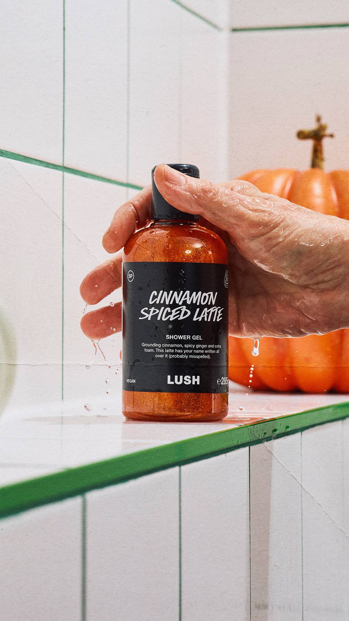 A wet hand reaches for a bottle of shimmering copper Lush Cinnamon Spiced Latte shower gel, resting on a white tiled ledge with green grout. A large orange pumpkin sits in the background.