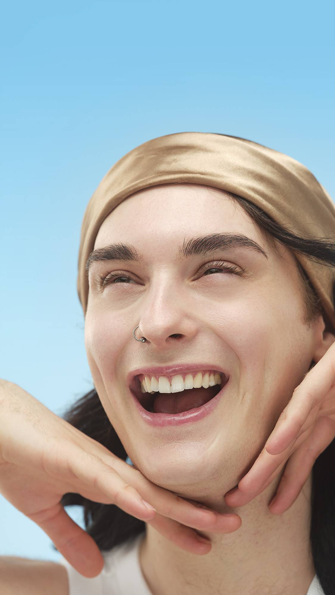 A smiling person with a gold headband and nose ring has their jawline framed by hands. They have defined brows and appear to be wearing black Circus Lashes ~ Mascara against a bright blue background.