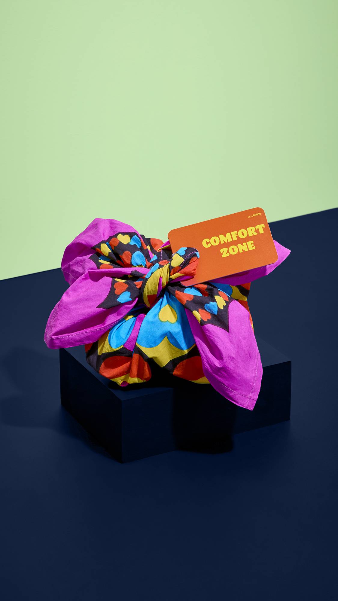 A vibrant gift wrapped in a colourful heart-patterned pink knot-wrap with an orange tag reading "COMFORT ZONE", displayed on a dark blue pedestal against a pale green and dark blue split studio backdrop.