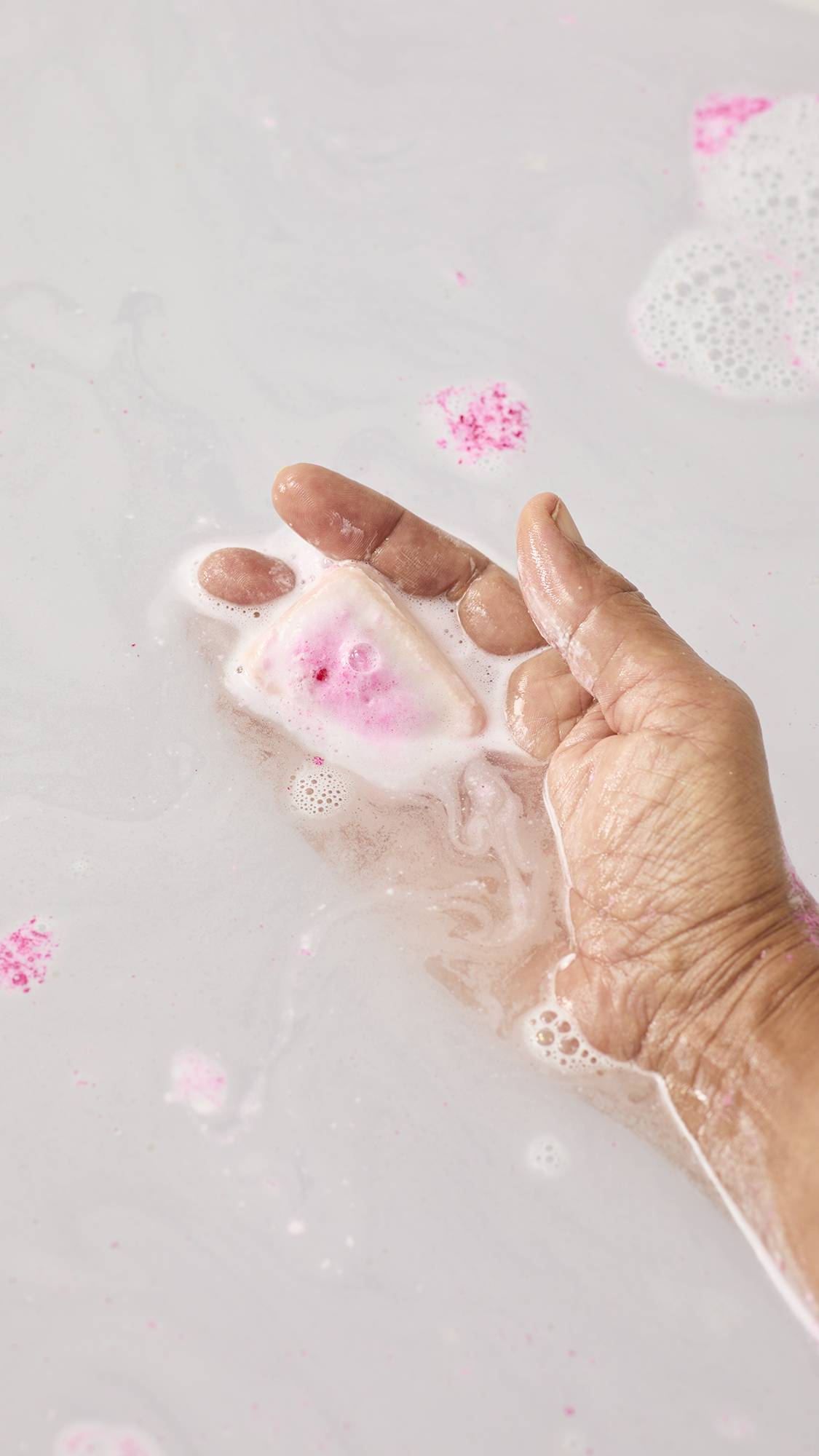 A hand holds the melting "Creamy Candy" bath melt in the bathwater, which has turned a milky white texture. Small pink flecks float on the surface as the bar dissolves, releasing its moisturizers into the water.