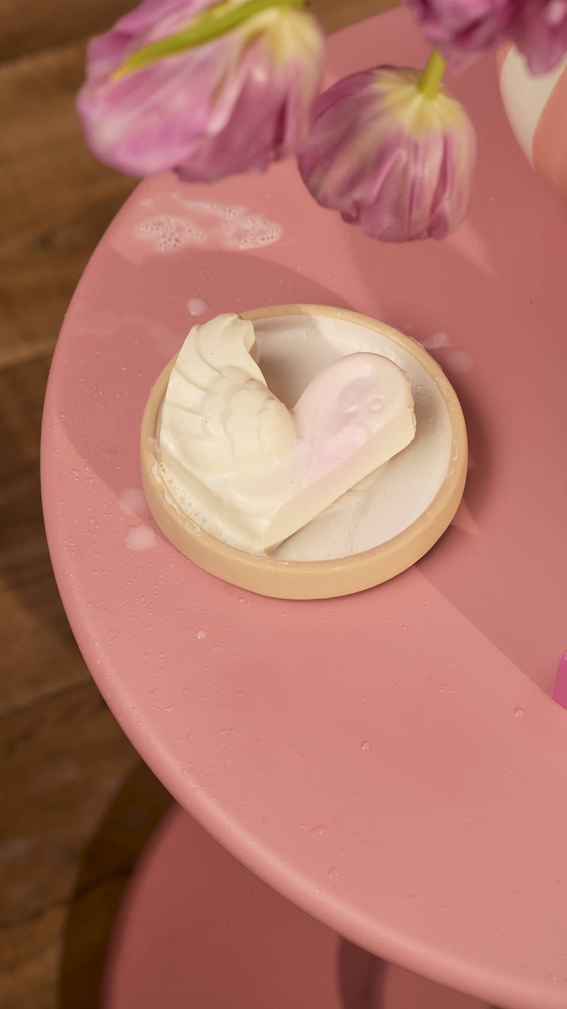 The white, Cygnet Ring single swan-shaped soap rests in a small, round ceramic dish on a pink table. The product is slightly wet with bubbles at the base, and soft pink flowers are blurred in the upper background.