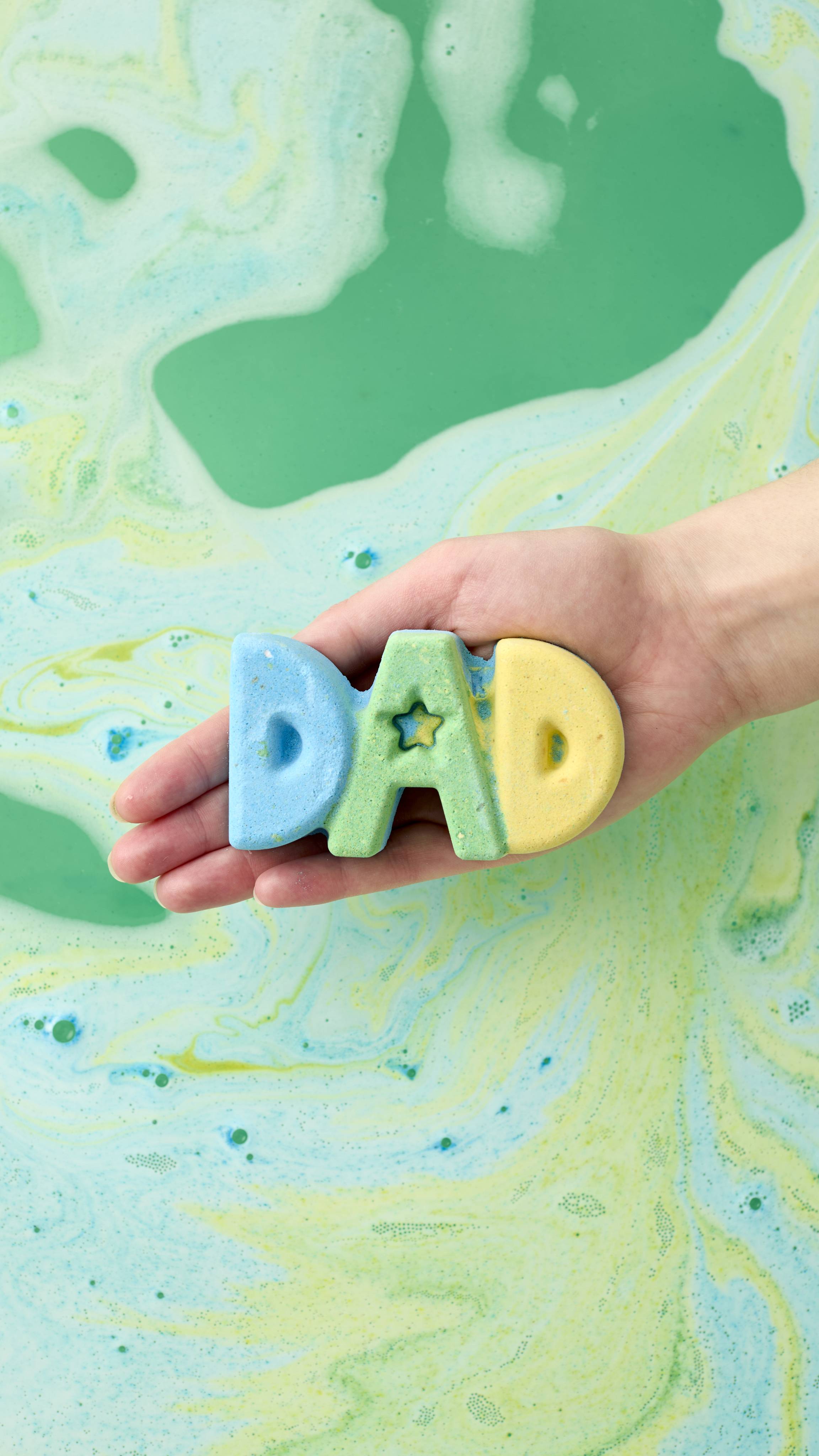A model's hand holds the blue, green, and yellow "DAD" bath bomb in an open palm over a bathtub. The water below is a vibrant, swirling mix of green, yellow, and blue foam, creating a marbled effect as the product begins to dissolve.