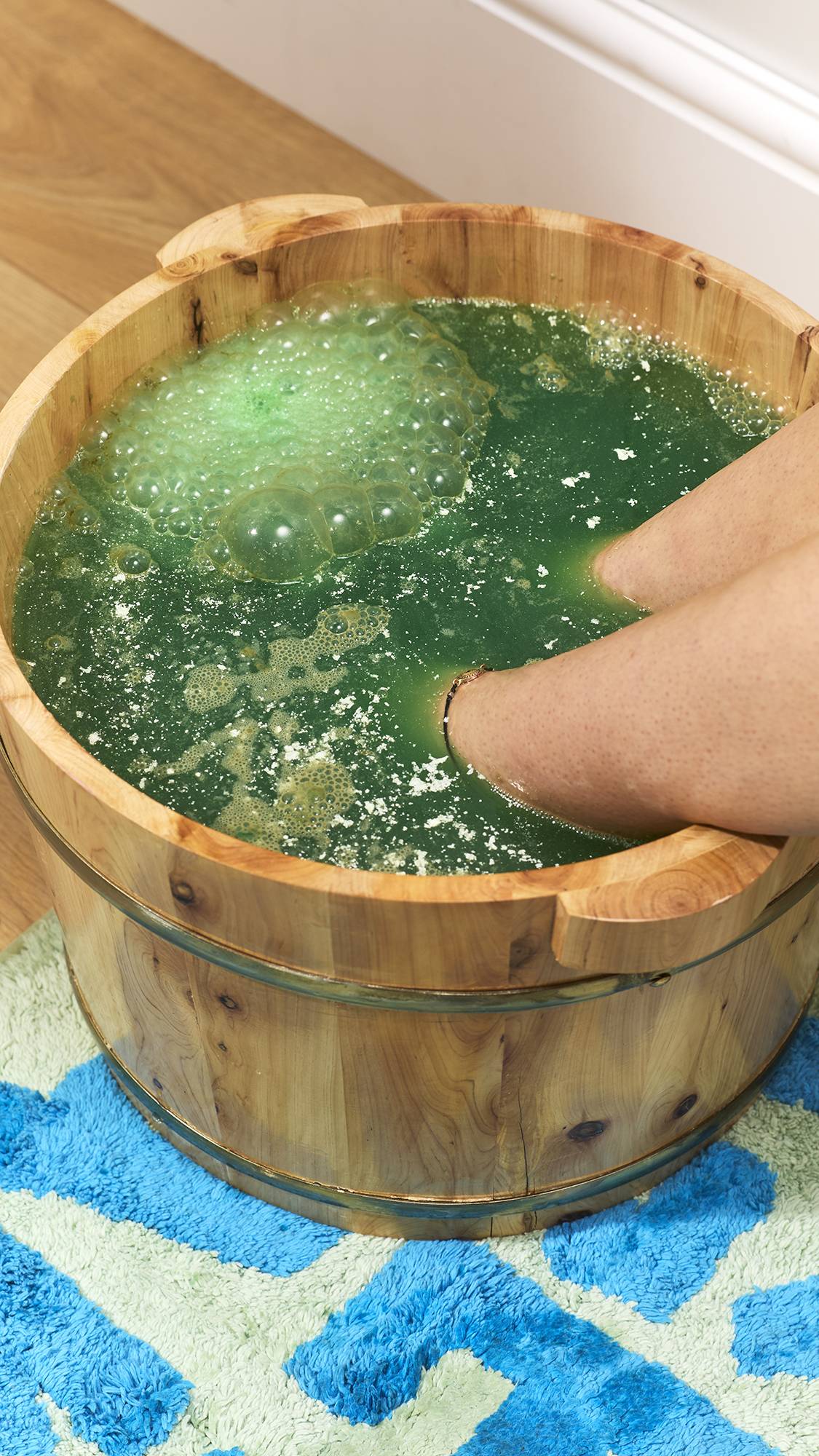 The image shows feet soaking in a wooden tub filled with dark green, sparkling bathwater releasing a cluster of light green bubbles and fine white shimmer, set on a blue patterned bathmat.