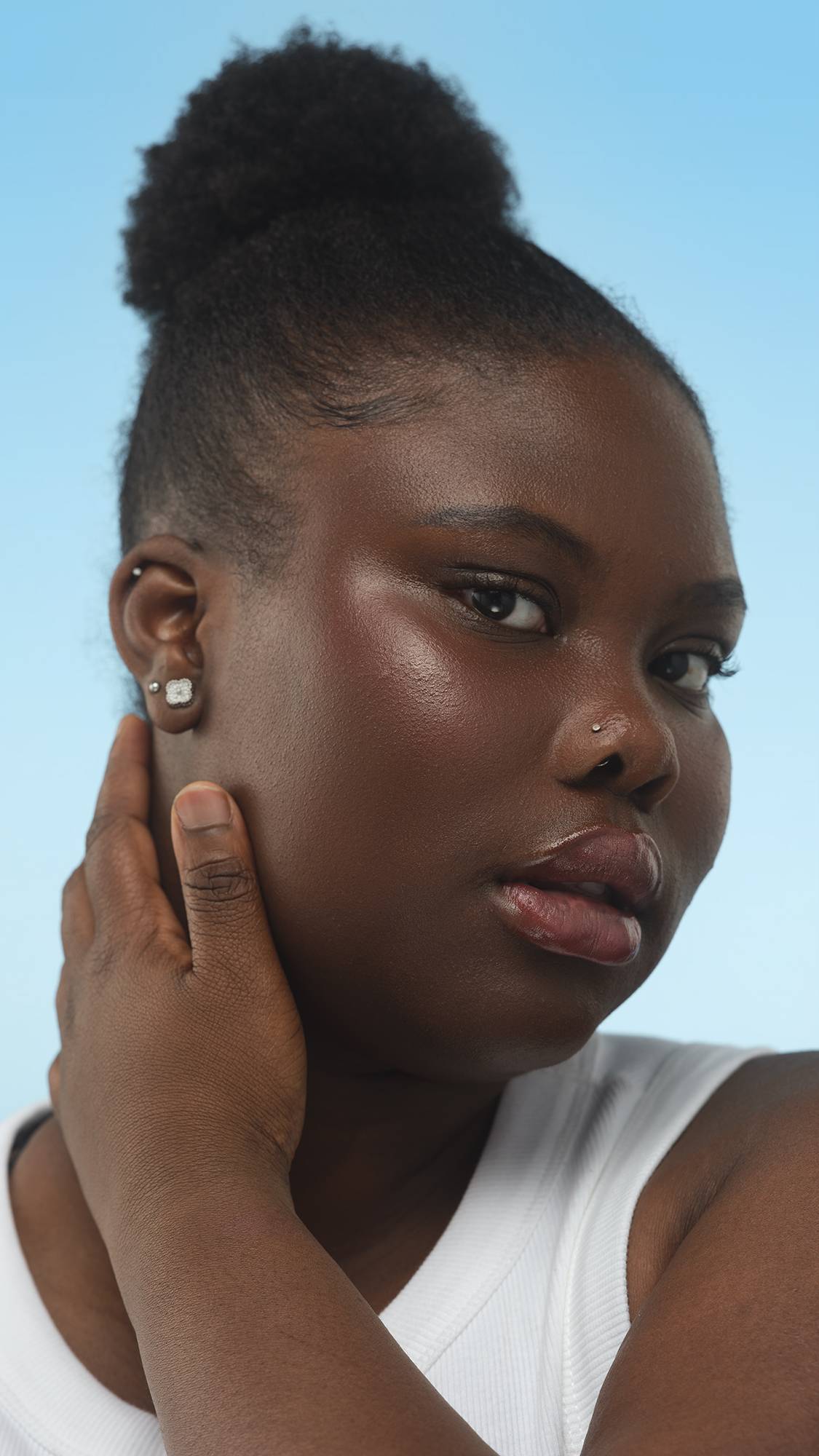 A close-up vertical shot of a person with deep skin, looking at the camera. Their dark hair is in a bun, and a subtle sheen from the Dark Cool Beauty Balm highlights their cheek as their hand rests near their ear, set against a bright blue background