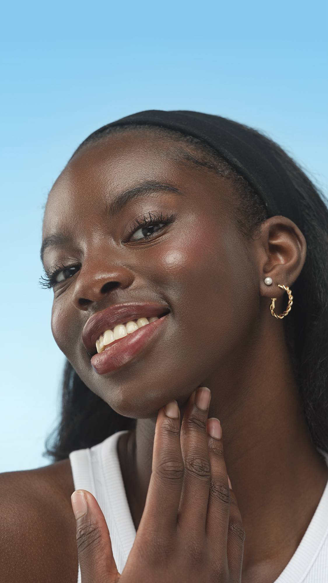 A close-up vertical portrait of a smiling person with deep skin, wearing a black headband and a white top. They wear the Dark Neutral Beauty Balm product and have gold hoop and pearl earrings, set against a bright, gradient blue background.