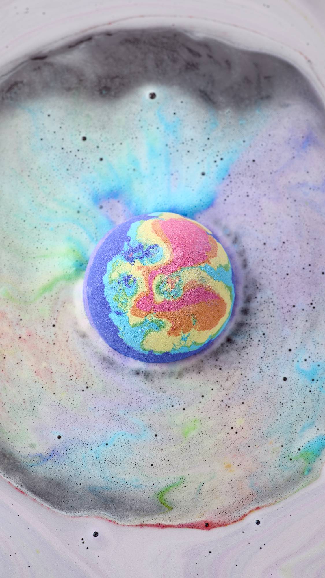 An aerial view of a vibrant, multi-coloured "Diversity" bath bomb dissolving in water. Swirls of neon pink, yellow, blue, and purple foam radiate from the centre, mixing with dark grey and pale lavender bubbles to create a cosmic, marbled effect.
