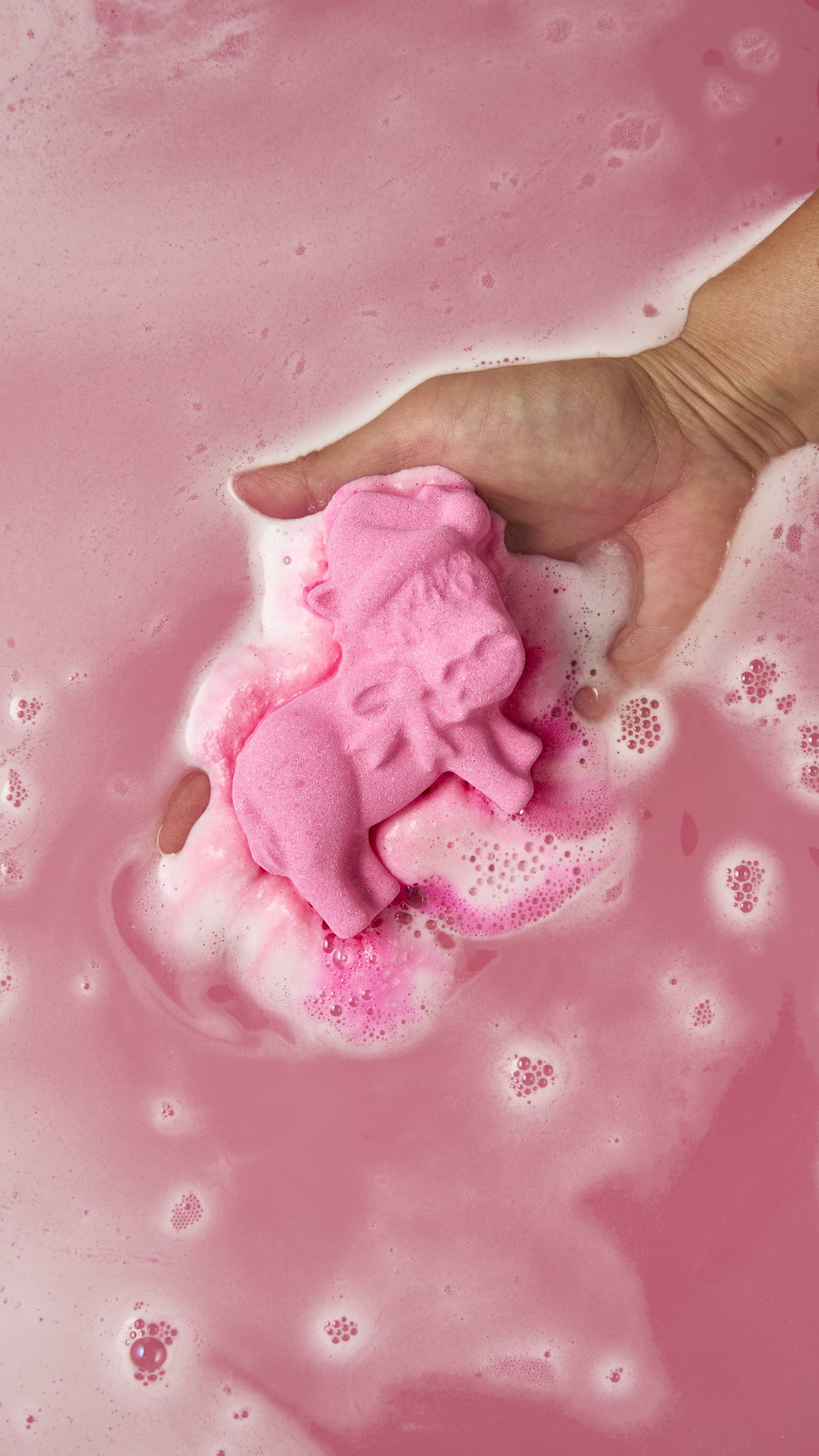 An aerial view image showing a hand gently placing a pink donkey-shaped bath bomb into pink water as the product fizzes, giving off ripples of deep pink foam.