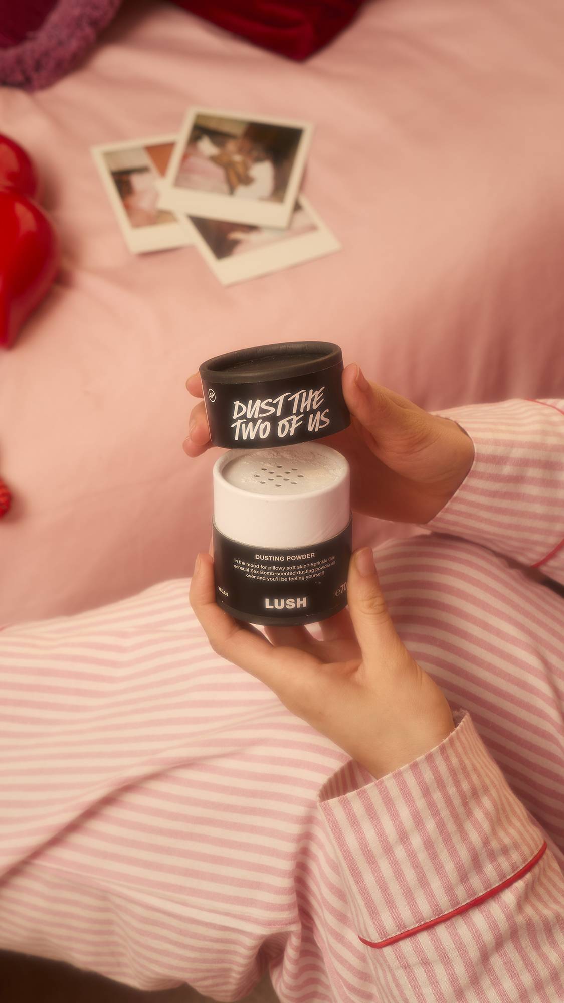 A person wearing pink and white striped pajamas holds a pot of Dust The Two Of Us dusting powder, lifting the lid to reveal the sifter top against a backdrop of pink sheets and polaroid photos.