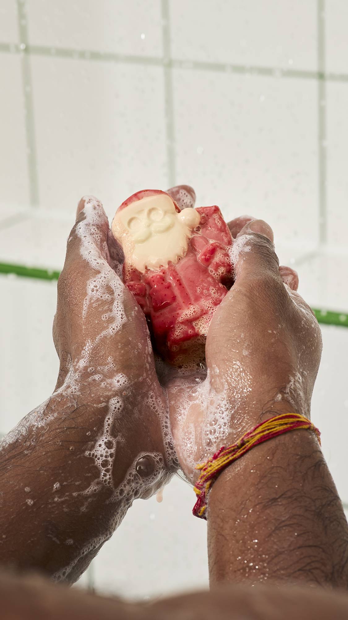 Hands hold and lather a red and cream Santa Claus-shaped soap bar in the shower, building up white bubbly suds across wet palms and wrists against a white tiled wall background.