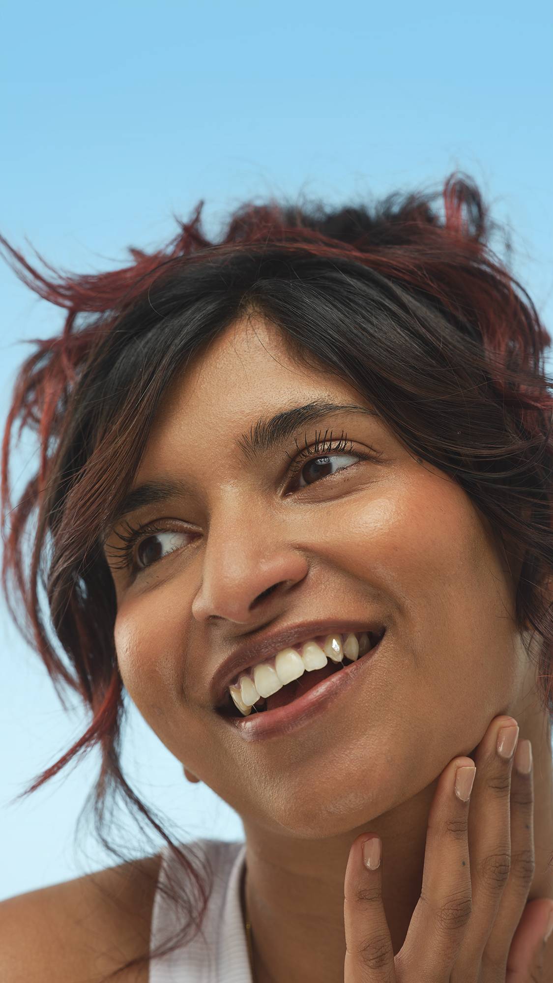 A person with dark hair with red highlights smiles and looks up and to the side, lightly touching their jawline. They have defined, lifted lashes coated with a black mascara, consistent with Eyes Right ~ Mascara, against a bright blue background.