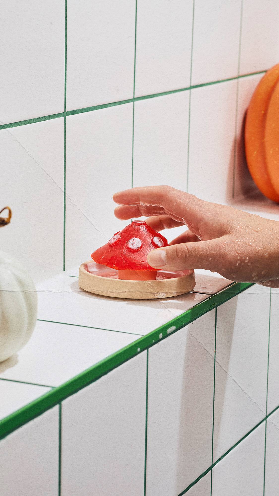 A wet hand reaches for a toadstool-shaped red soap resting on a small dish along a tiled bathroom ledge with green grout, flanked by ornamental pumpkins.