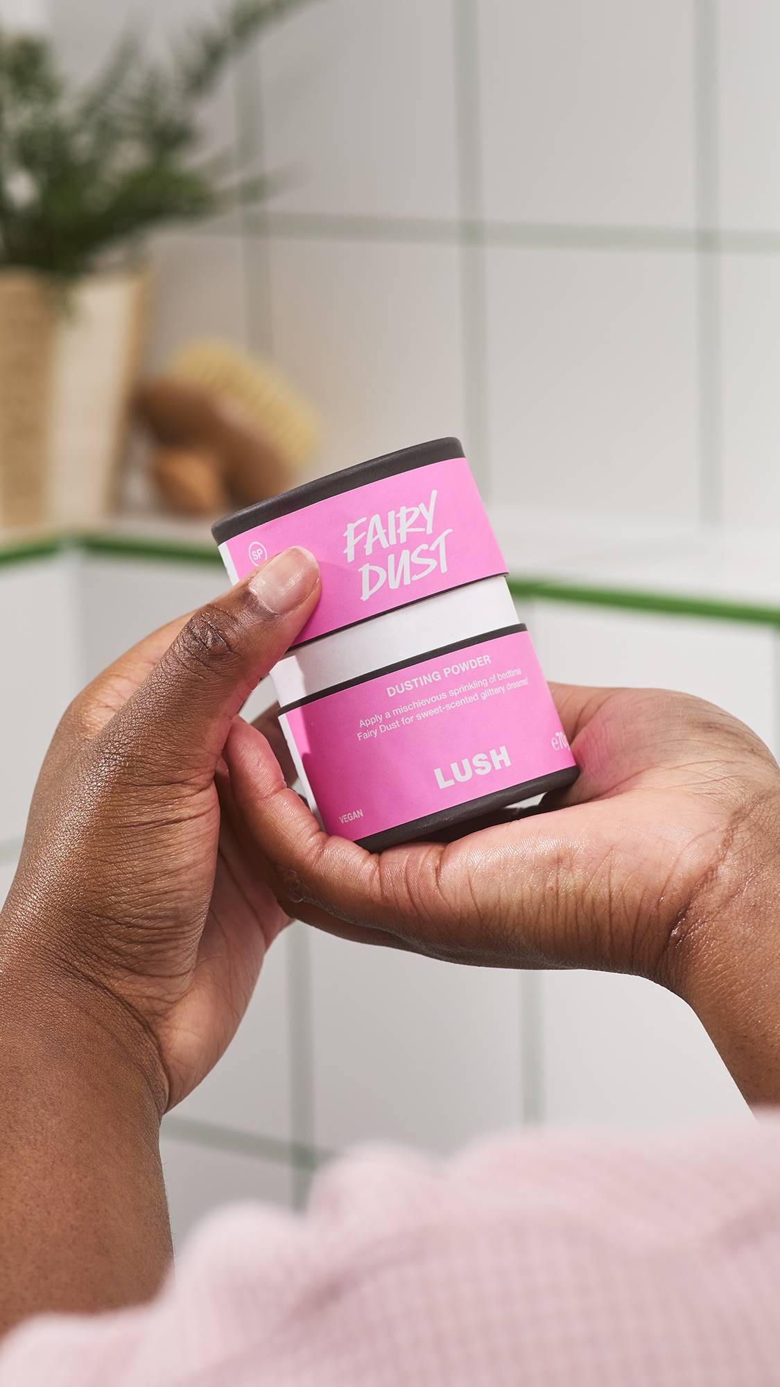 Hands hold open a pink and black container of Lush Fairy Dust dusting powder, set against a white tiled bathroom background with potted plants.