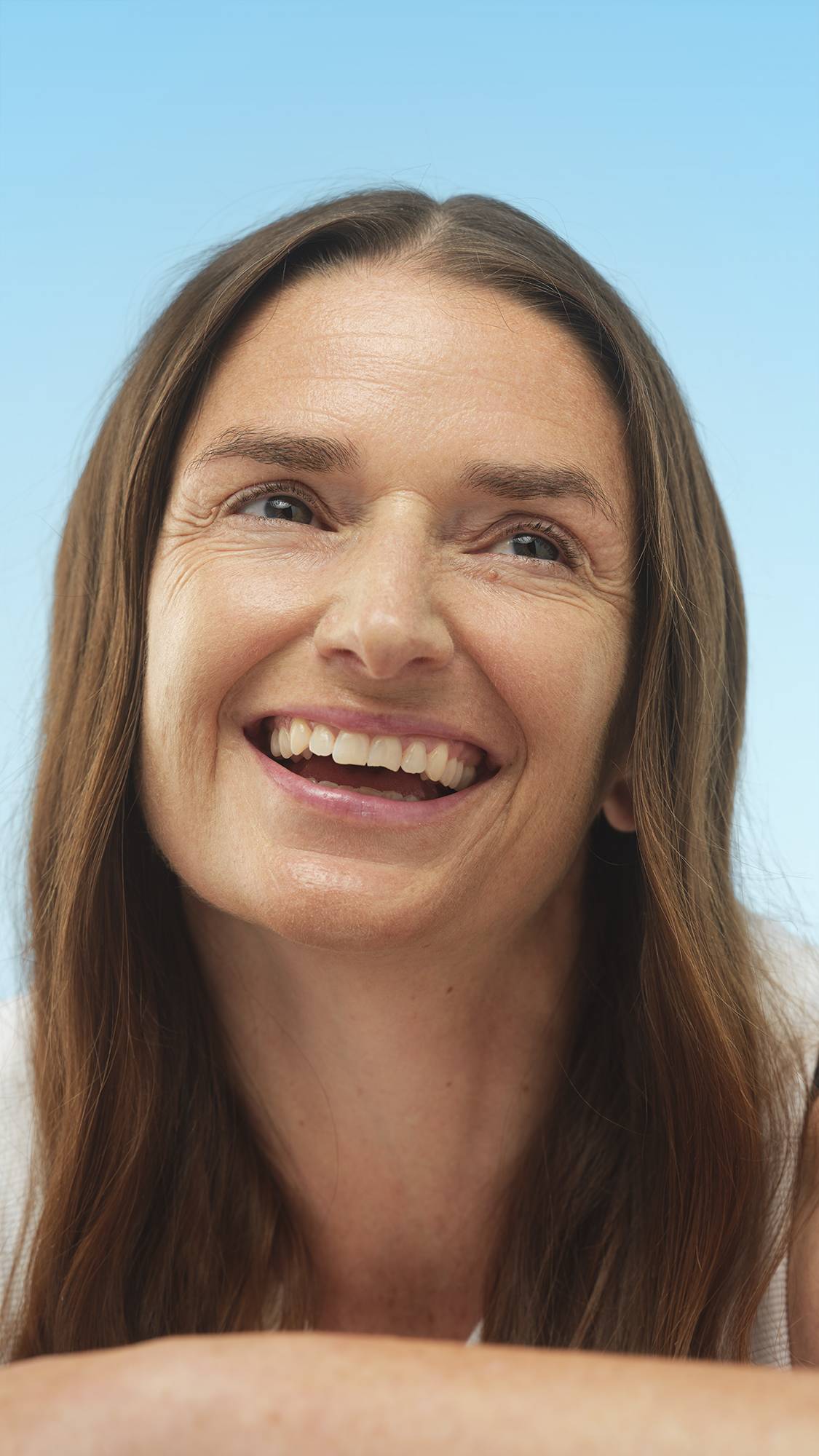 A close-up vertical portrait of a broadly smiling person with long brown hair, looking up. Their face has a natural, even tone, prepped with the Feeling Greener primer, set against a bright blue background.