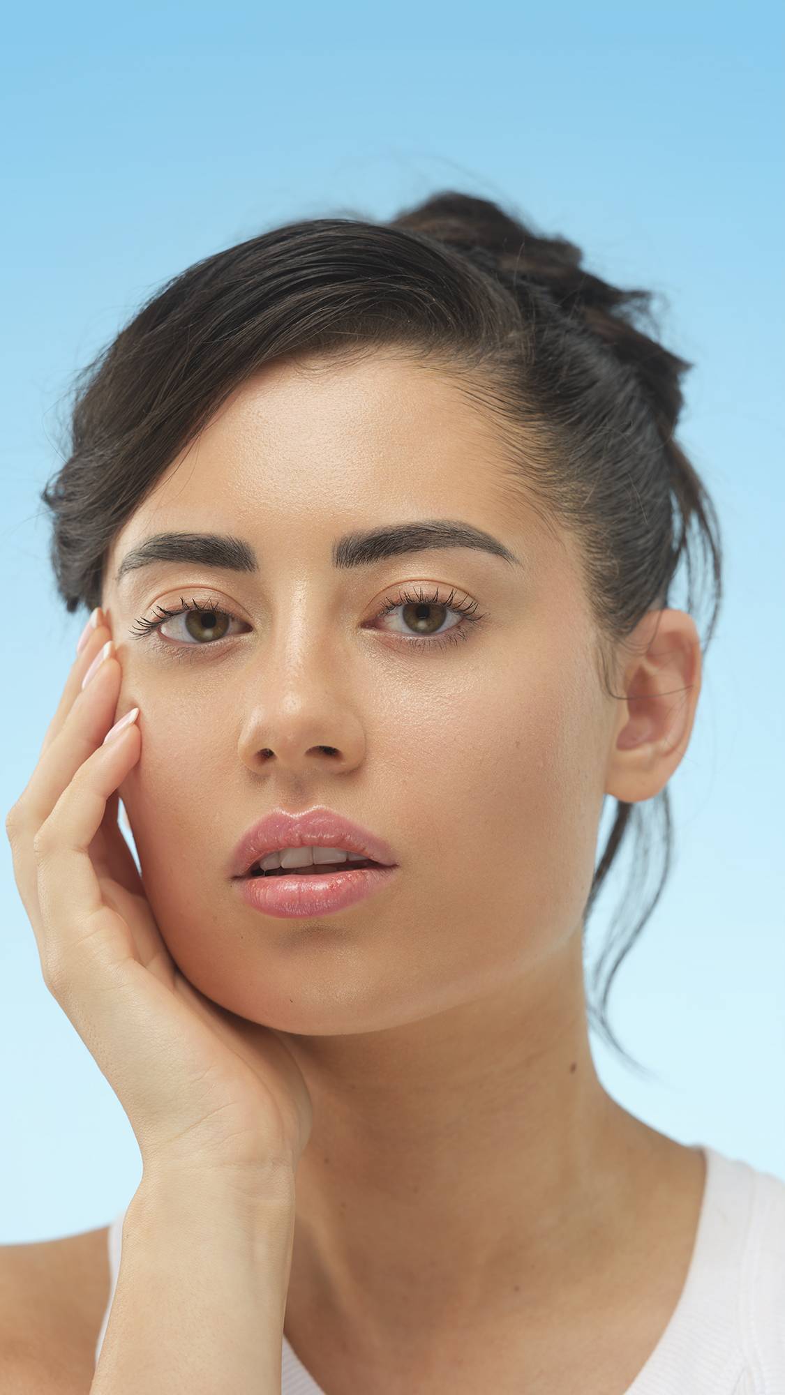 A close-up portrait of a person with medium skin and dark hair, looking directly at the camera with their hand on their cheek. Their skin has a noticeable glow, likely from the Feeling Younger Skin tint, set against a bright blue background.