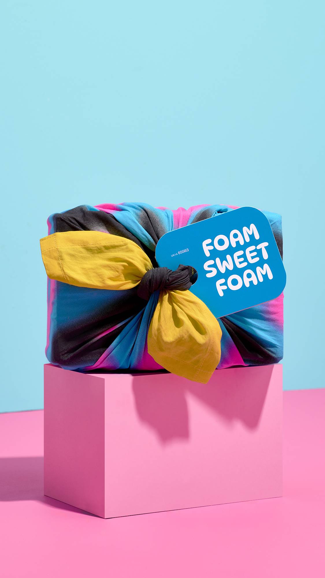 A gift wrapped in vibrant knot-wrap fabric with yellow tied ends sits on a pink block, tagged with a blue label reading "FOAM SWEET FOAM" against a sky-blue background.