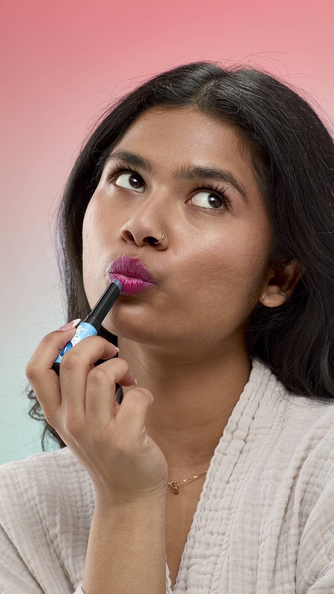 A person with long dark hair applies the shimmering, deep berry-purple Frankenstain lip stain to their pouted lips. They are holding the light blue product tube and looking upwards against a soft pink-to-blue gradient background.