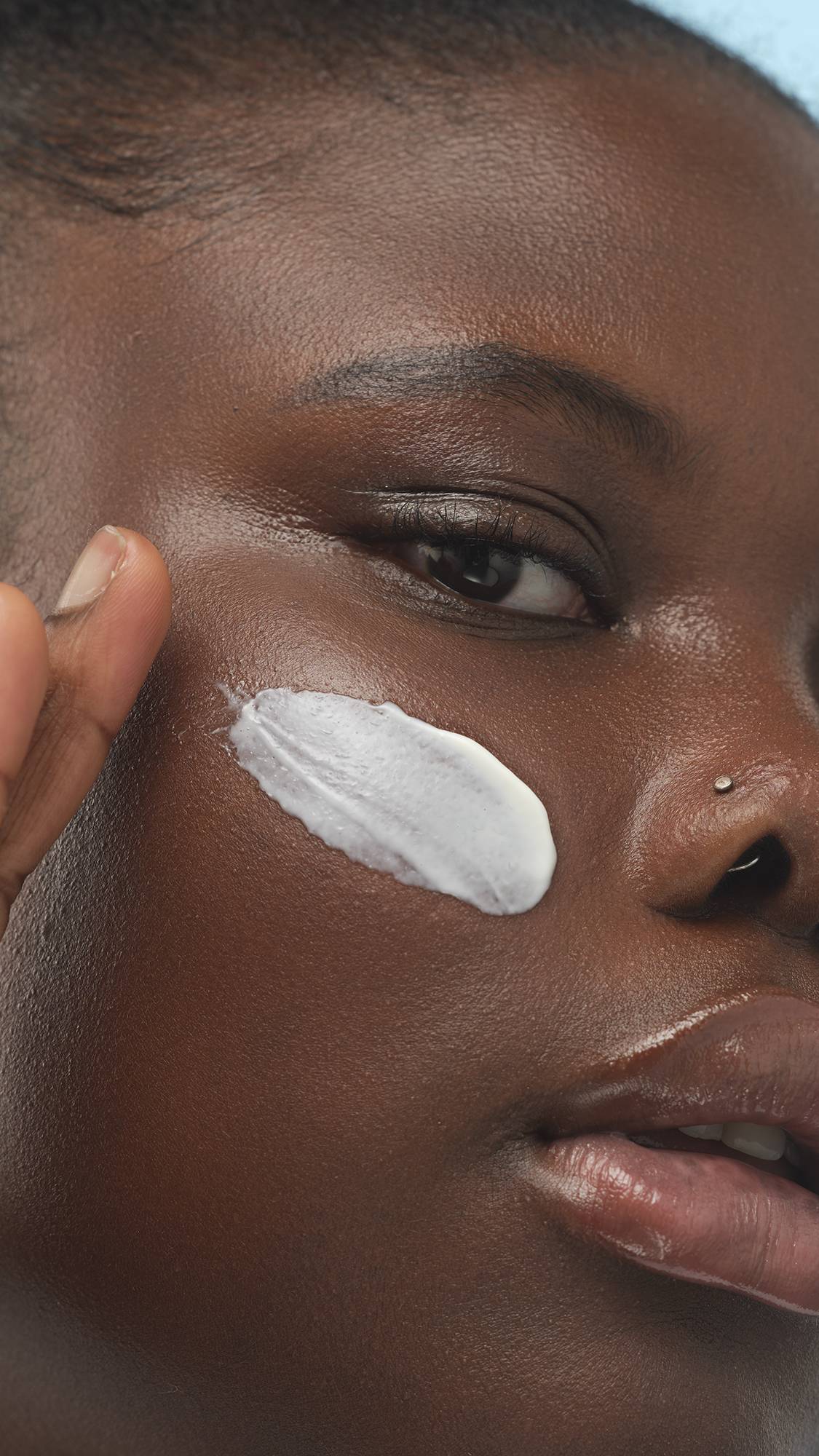 A close-up vertical shot of a person with deep skin, applying a creamy white swatch of set primer to their cheek with a finger. The skin has a healthy sheen, and the person has a nose stud.