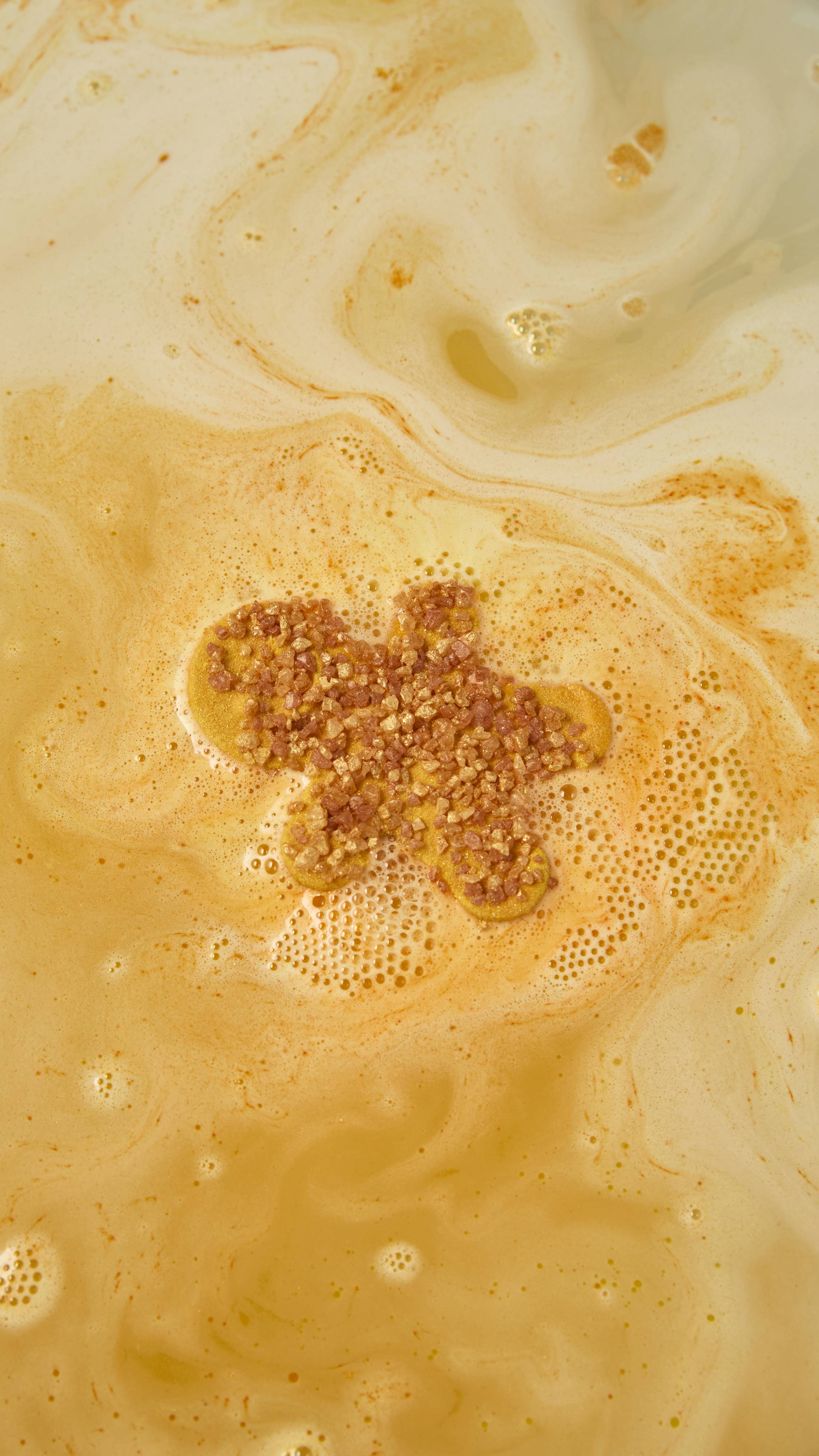 A gingerbread man bath bomb dissolves in bathwater, releasing a shimmering pool of golden-yellow foam around its crystal-topped body as soft, creamy waves spread across the warm amber surface.