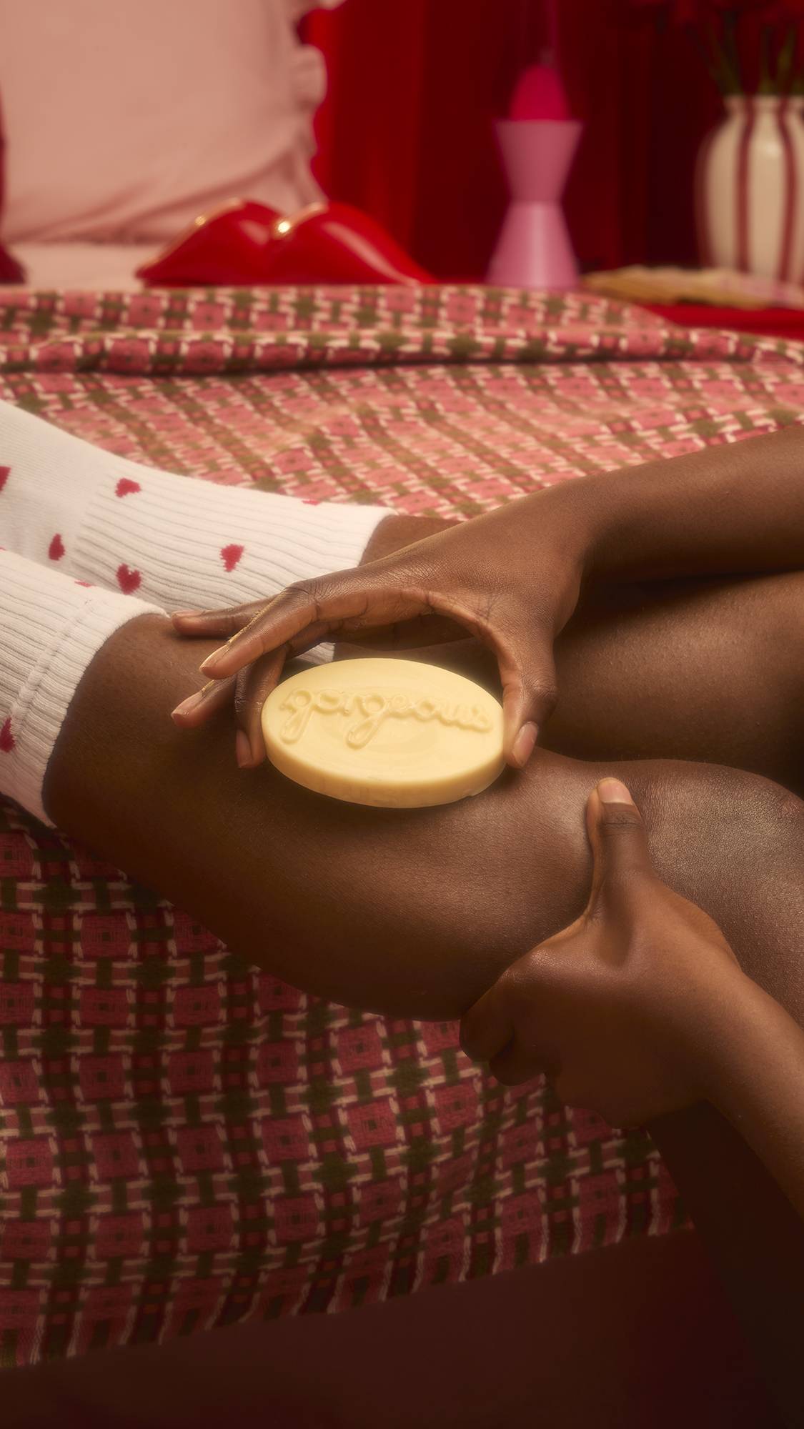 A cream-coloured, oval-shaped massage bar with the word "gorgeous" embossed across its top surface is held against a person's leg. The model is sitting on a patterned red and white bedspread, wearing white socks with small red hearts.