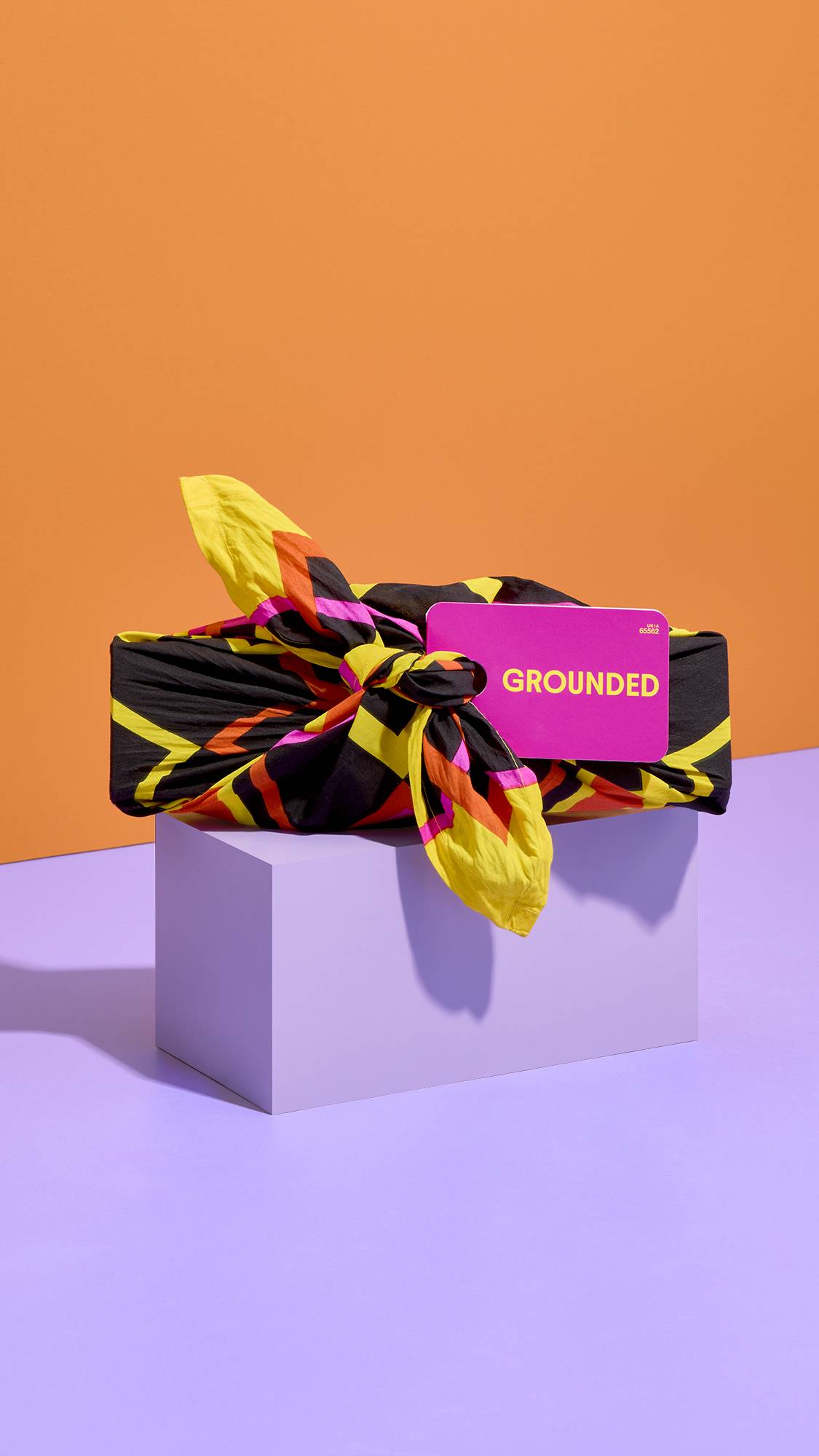 A LUSH knot-wrapped gift patterned in black, yellow, orange, and pink sits on a lilac plinth, featuring a pink tag that reads "GROUNDED" against a split backdrop of warm orange and purple.