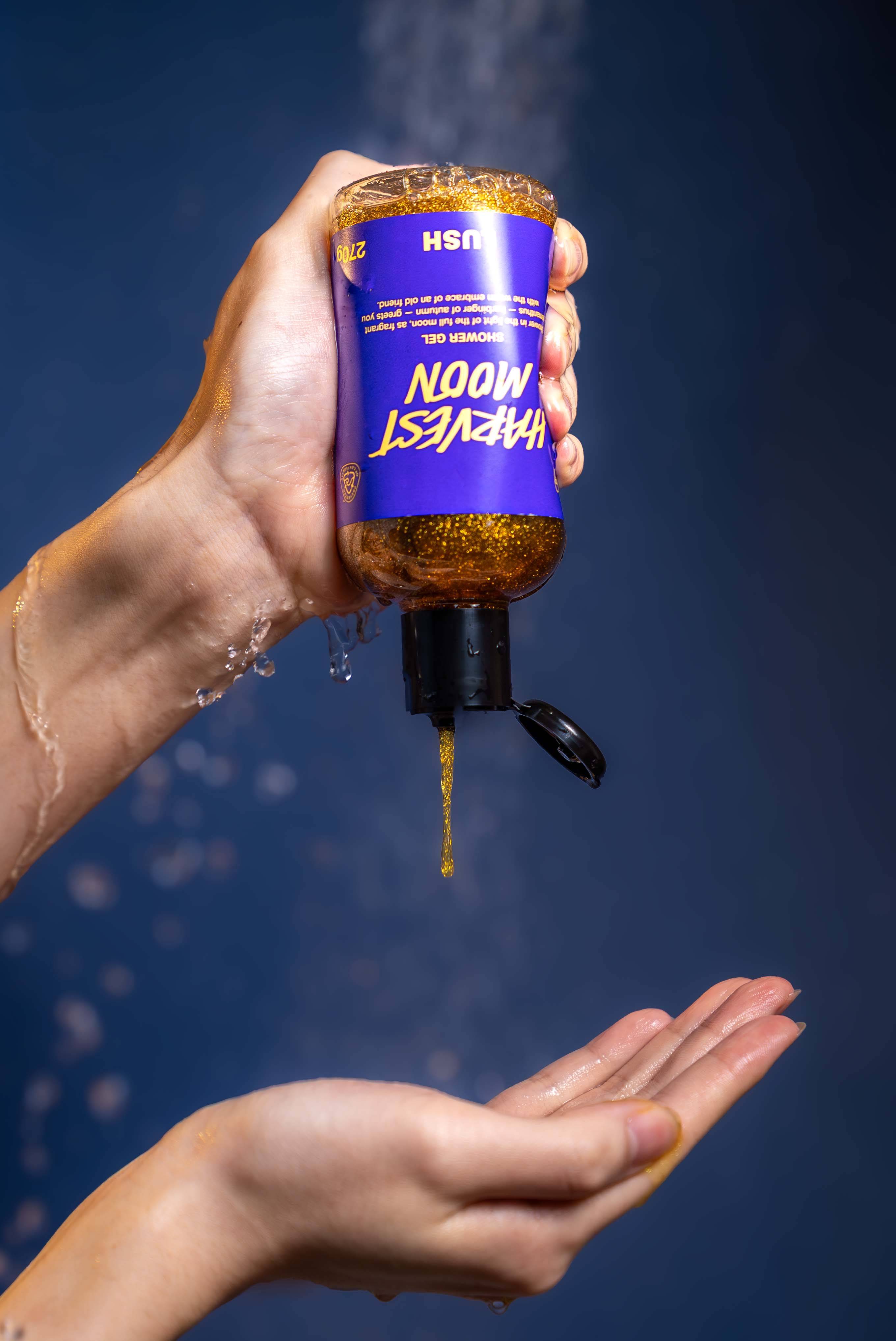 A hand holds an upside-down bottle of Lush "Harvest Moon" shower gel, squeezing a stream of glittery golden liquid into an open palm below. The background is a deep blue with blurry water droplets spraying downwards.