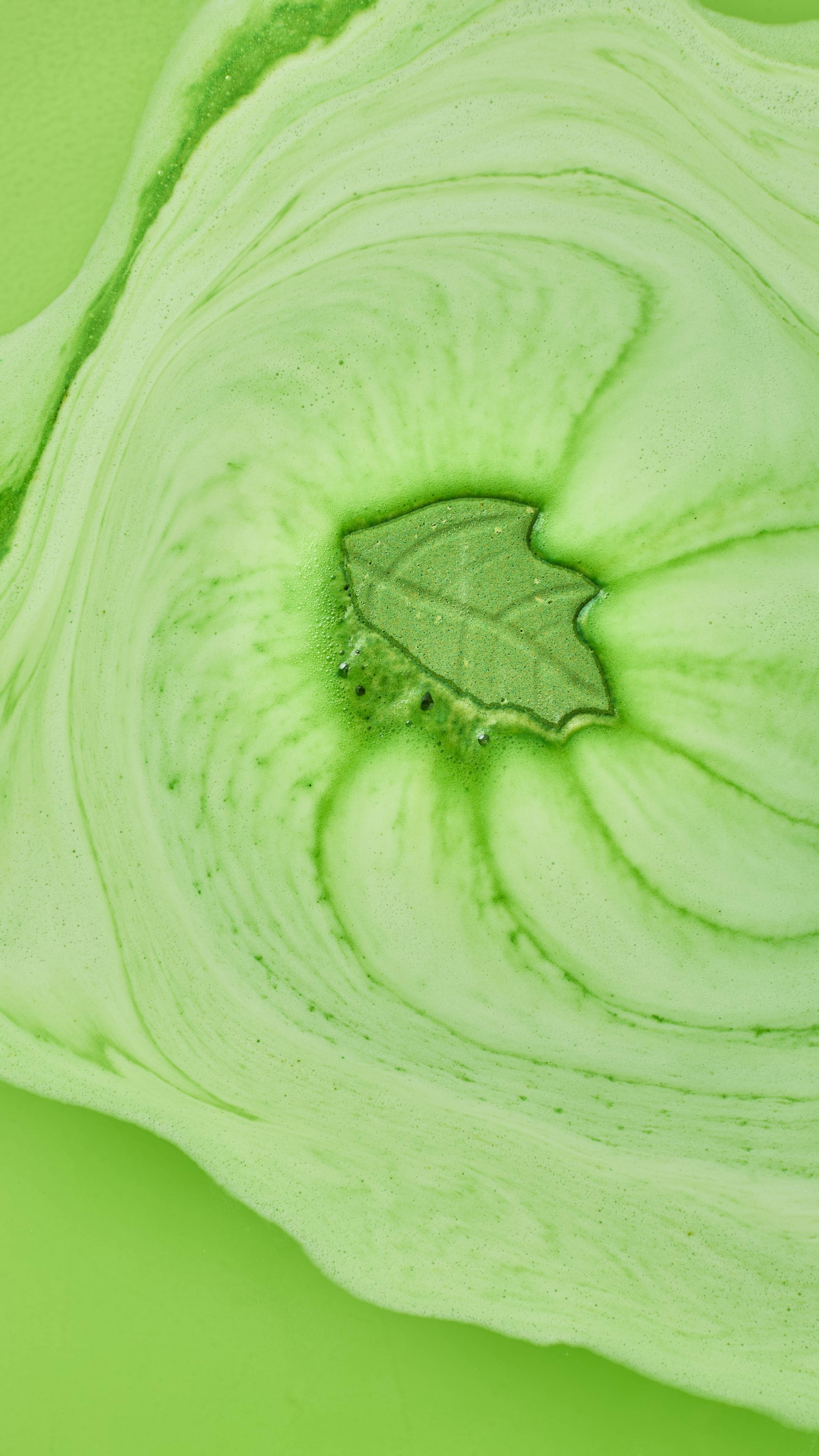A top-down view of a small green leaf-shaped bath bomb dissolving in the centre of soft, pale green foam swirled with darker green streams across bright lime bathwater.