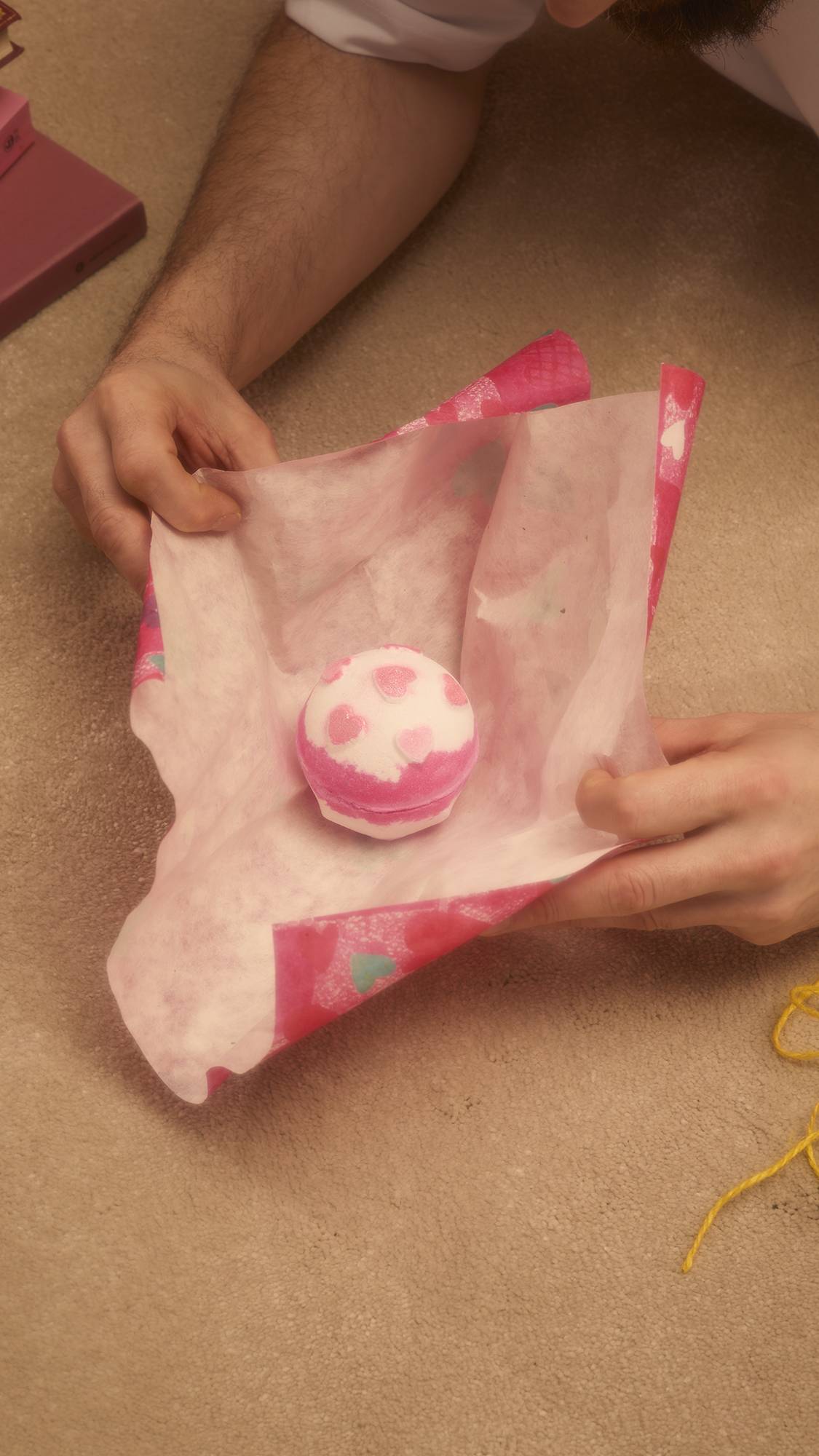 Hands hold open the pink Hearts Lokta Paper to reveal a white and pink bath bomb decorated with pink hearts. The paper features a pattern of colourful hearts on the reverse side.