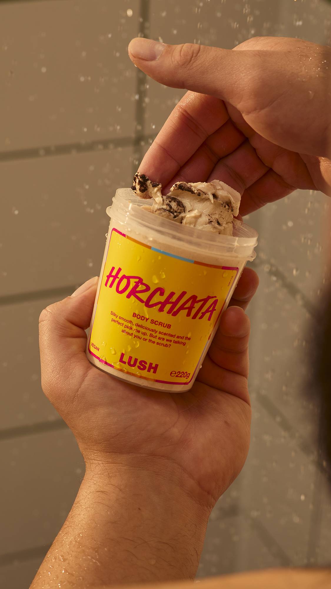 A person holds a pot of Lush Horchata body scrub in the shower, scooping out a portion of the thick, creamy, speckled paste with their fingers against a tiled wall.