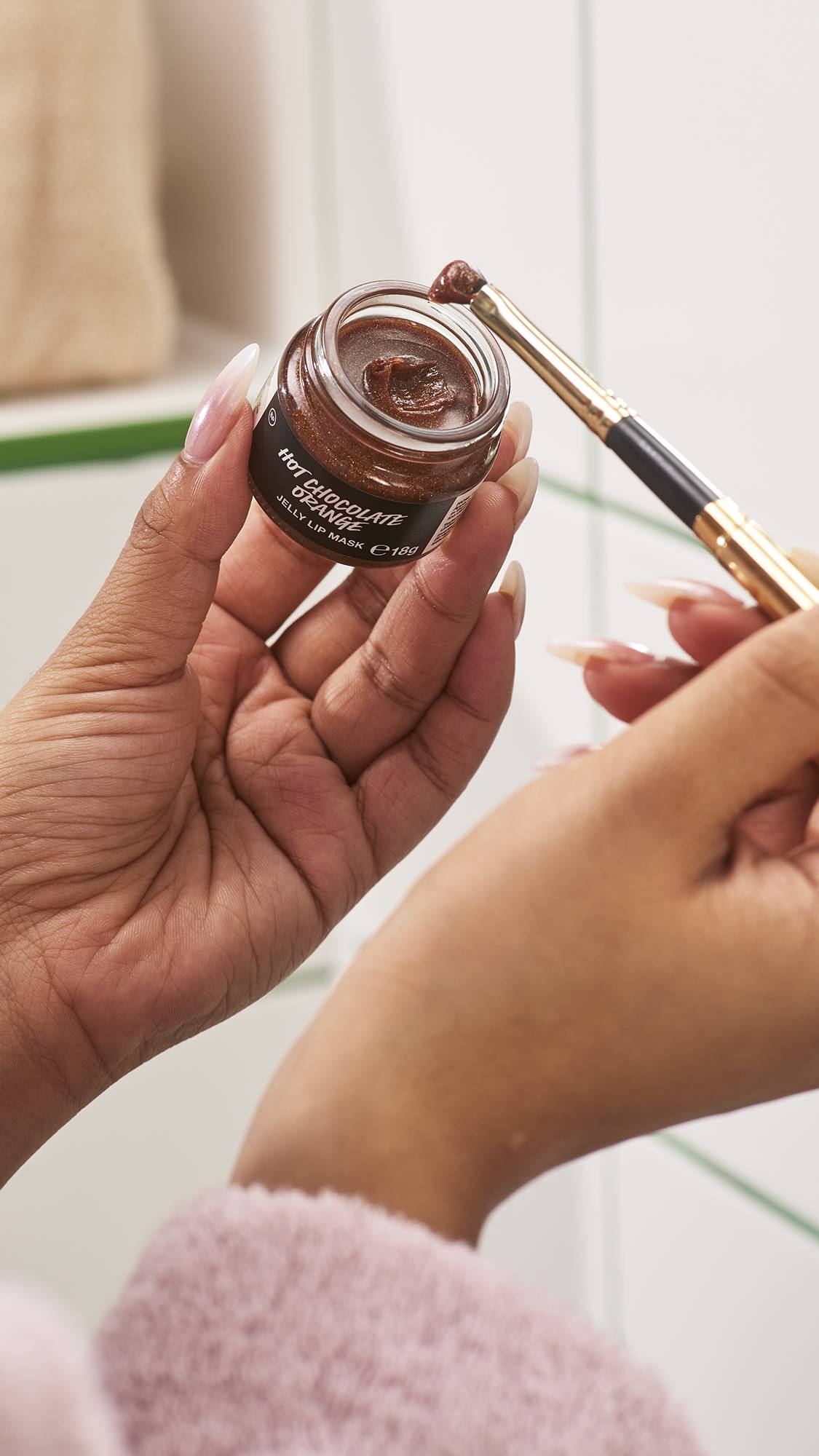 A person holds a small glass jar of Lush Hot Chocolate Orange jelly lip mask in one hand, using a small makeup brush in the other to scoop up the shimmering brown product, set against a white tiled bathroom wall background.