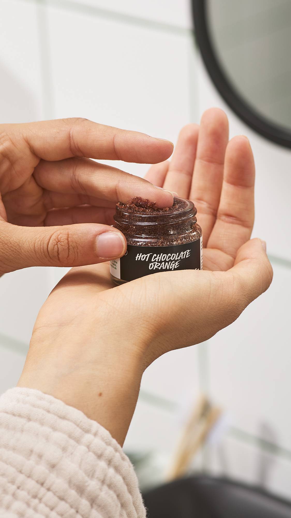 A person holds a small glass jar of Lush Hot Chocolate Orange lip scrub in one palm, dipping a index finger into the dark brown sugar scrub against a white tiled bathroom background.