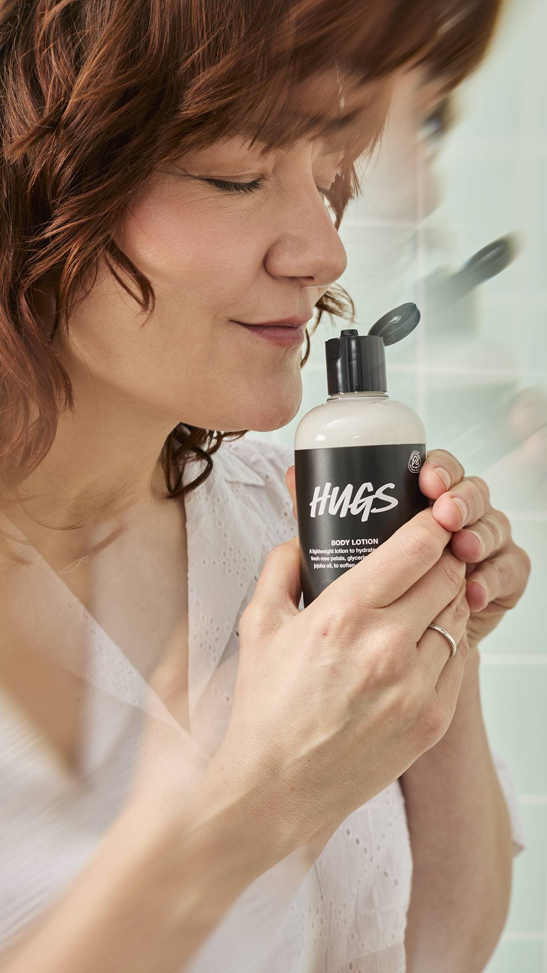 A model with auburn hair holds a bottle of Lush "Hugs" Body Lotion close to their face with a serene expression while smelling the scent. The background features blurred, light green tiles.