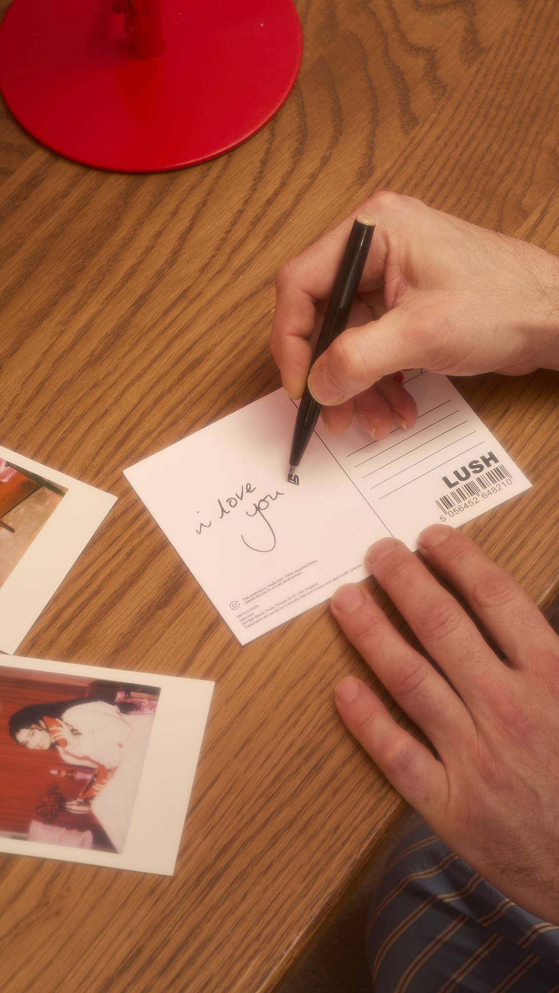 A person's hands write the message "i love you" in cursive on the back of a white Lush postcard using a black pen. Polaroid photos are scattered nearby on the wooden table surface.
