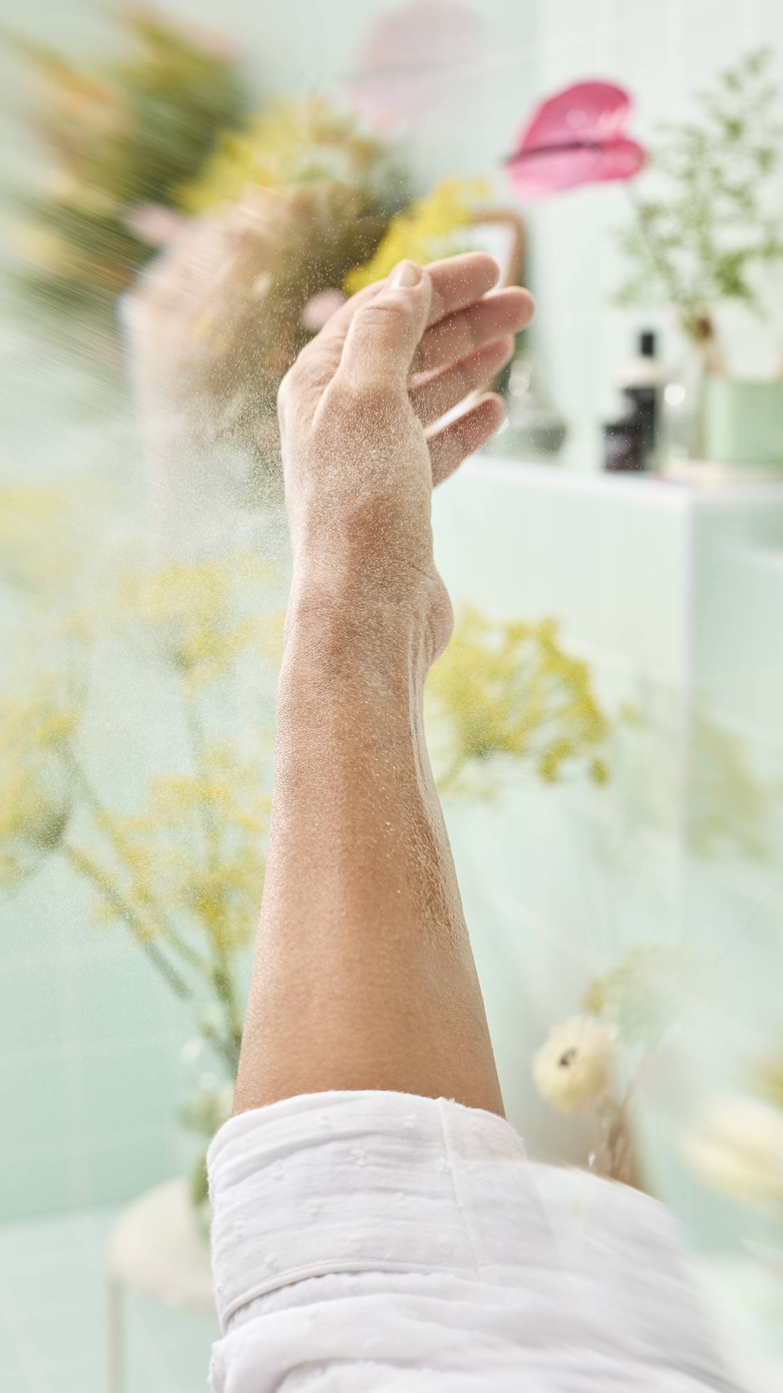 A close-up image showing a fine mist of "I Love You To Peaches" body spray being spritzed onto a model’s raised forearm. The background is a bright, soft-focus bathroom with pale green tiles and various colourful flowers.