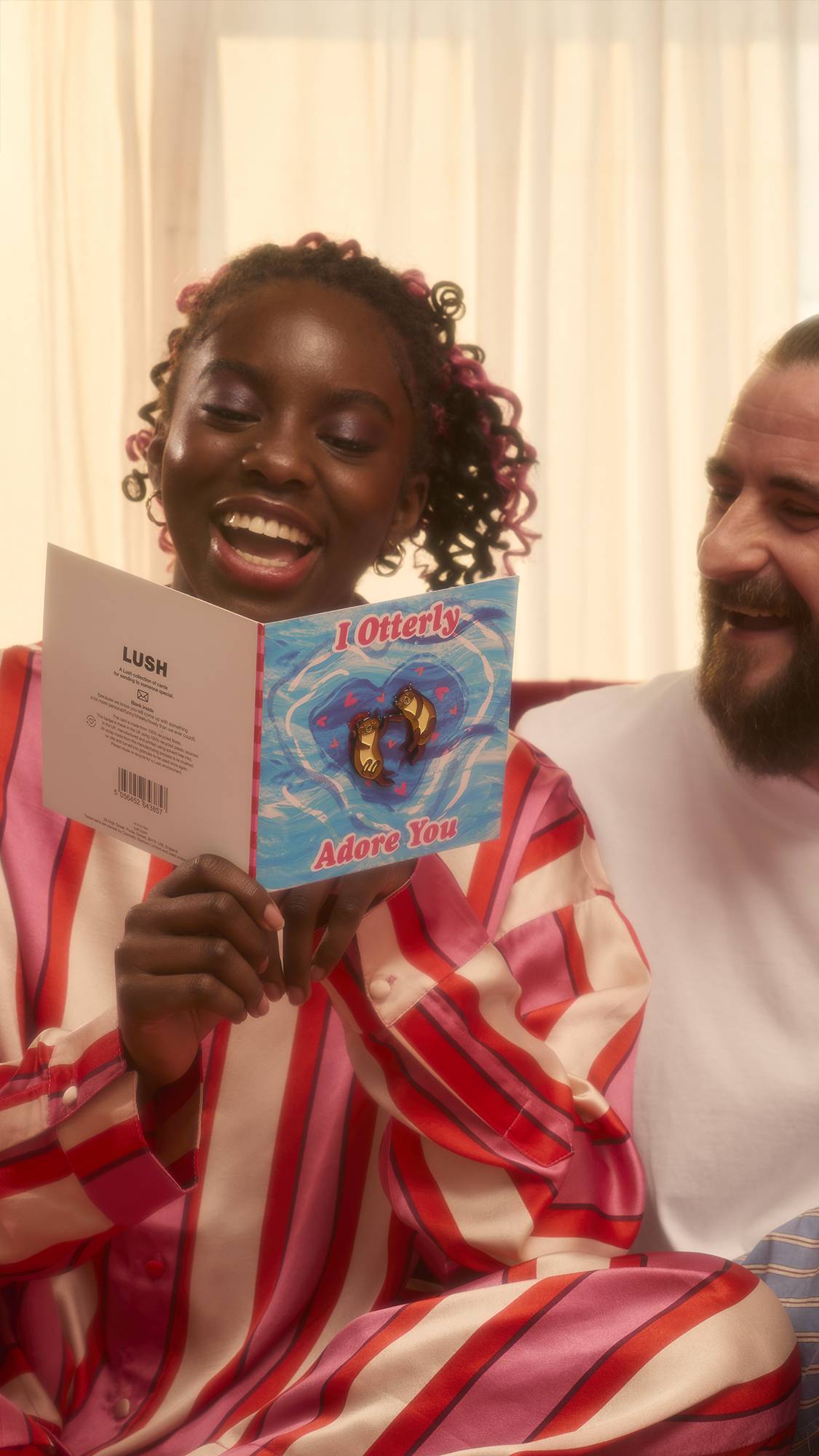 A smiling person wearing pink and red striped pajamas holds the open "I Otterly Adore You" card, sharing a joyful moment with a companion. The card features a blue water design with two otter pins attached.