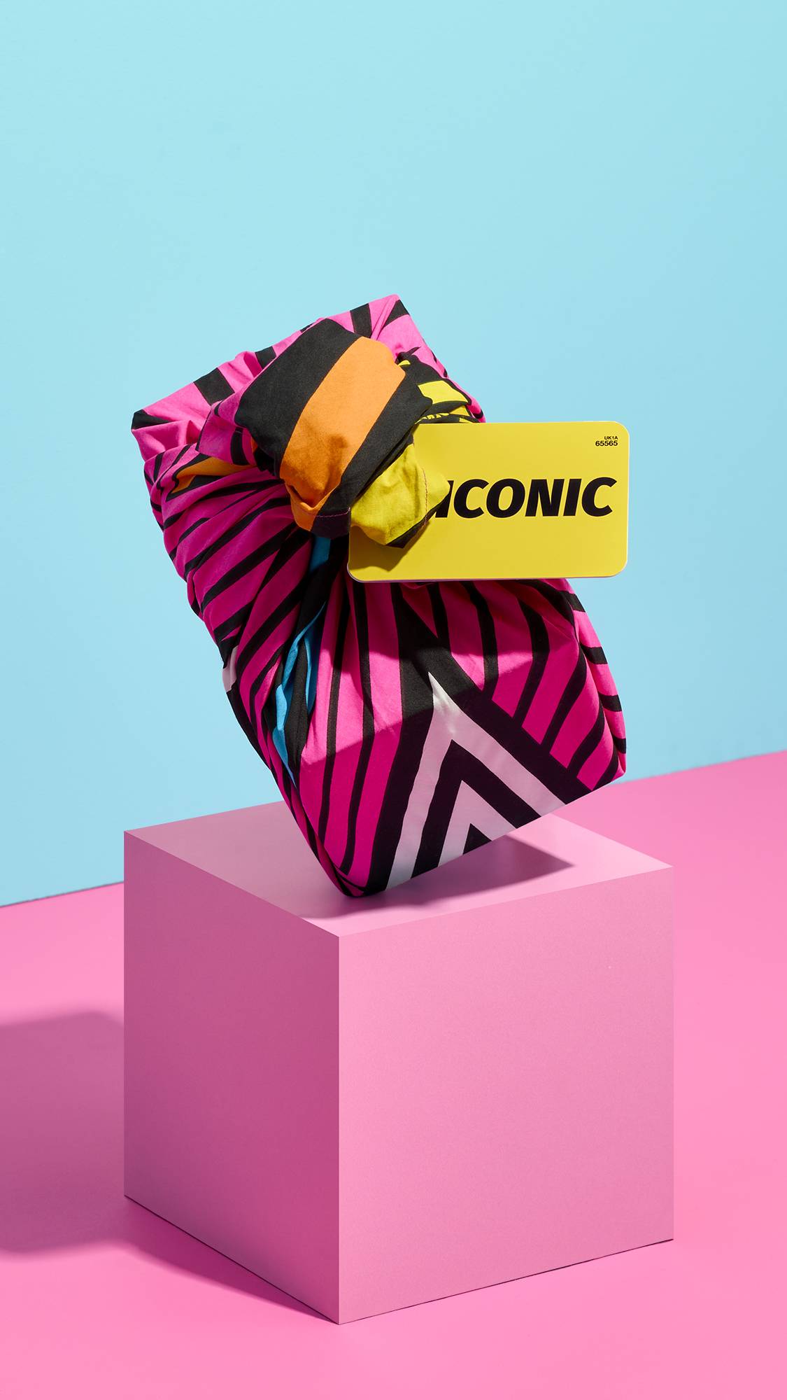 A knot-wrapped "ICONIC" gift set tied in a bright pink and black striped fabric balances at an angle atop a pastel pink block pedestal, set against a contrasting bright blue wall.