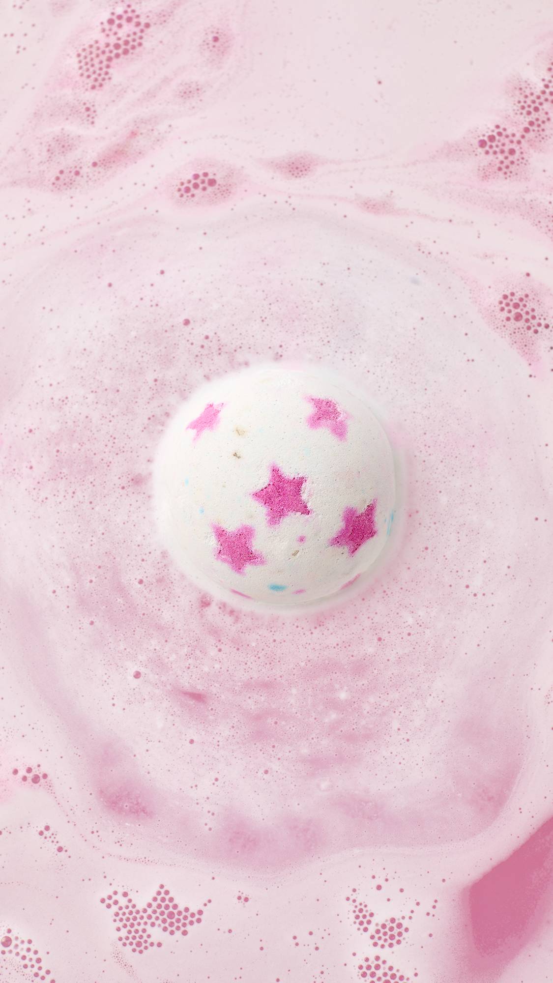 An aerial view of the dissolving Inclusion bath bomb. Thick, velvety swirls of bright pink, orange, and yellow foam ripple across the surface of the bathwater.