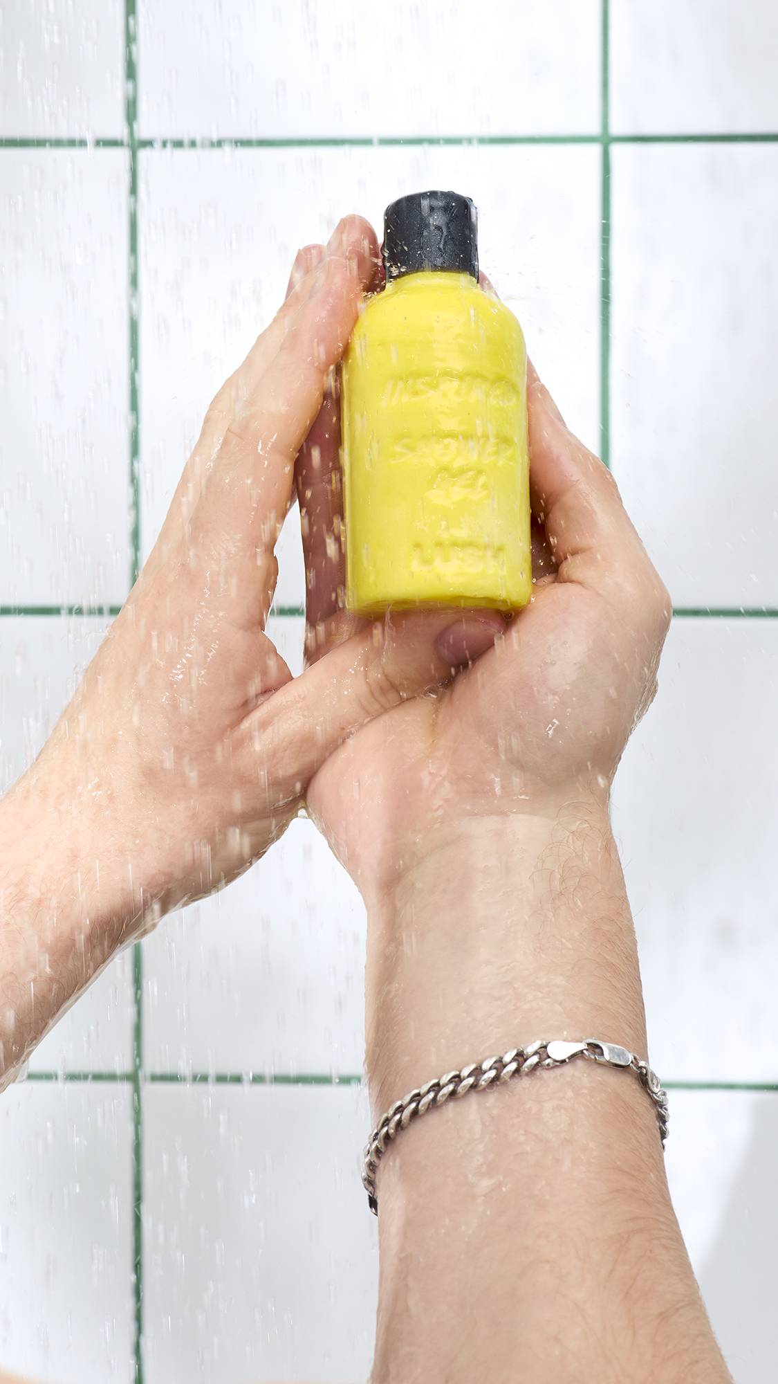 Wet hands hold a yellow, bottle-shaped "Inspire" solid shower gel under streaming water in the shower. A silver chain bracelet is visible on one wrist against a background of white tiles with green grouting.
