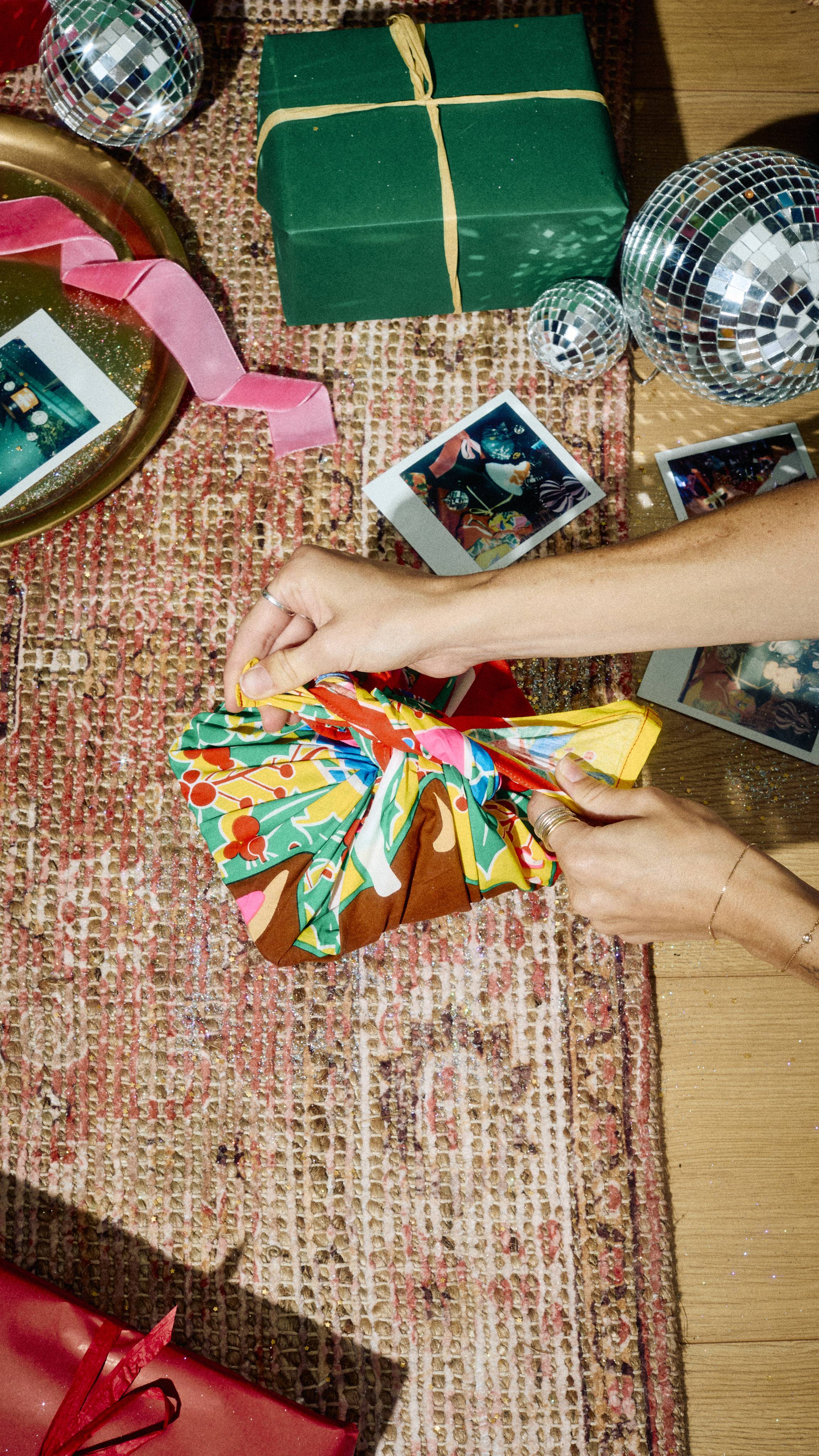 Hands tie a knot on a colourful reindeer-patterned Lush fabric Knot Wrap covering a gift, viewed from above on a patterned rug surrounded by wrapped presents, instant photos, and disco balls.