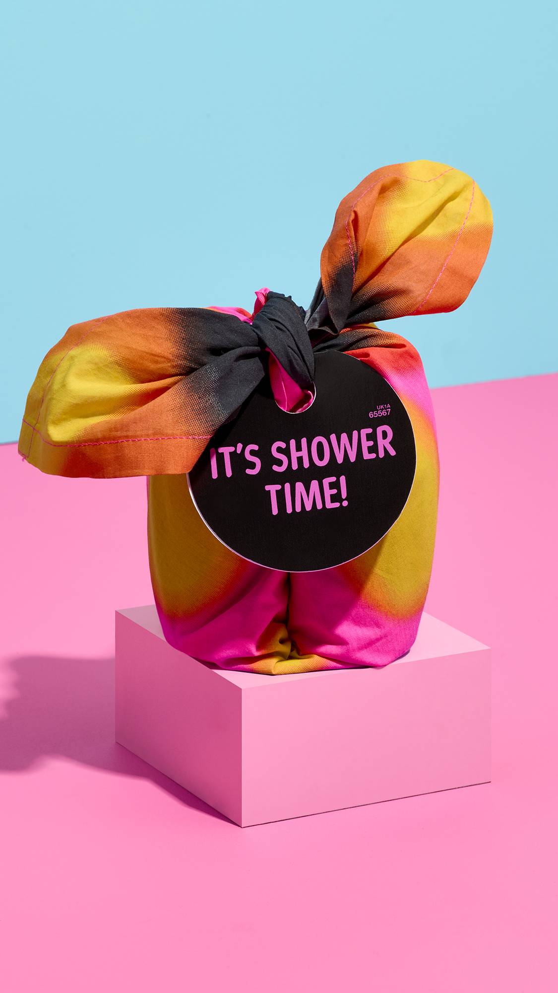 A tie-dye fabric-wrapped gift in neon pink, orange, and yellow sits on a light pink block, featuring a black circular tag reading "IT'S SHOWER TIME!" against a split background of pink and sky blue.