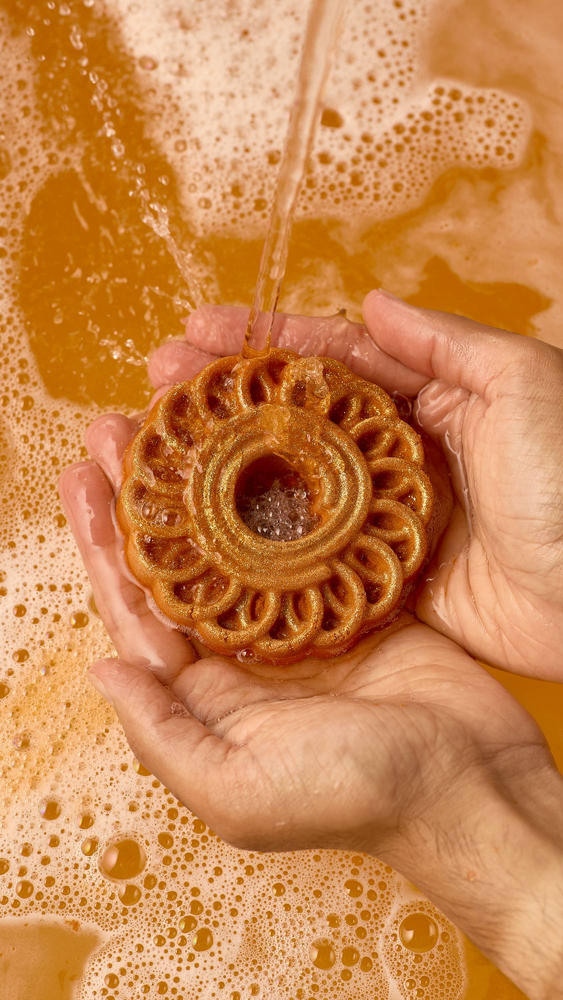 Two hands hold a round, golden-bronze shimmer Lush "Jalebi" bubble bar as running water pours directly over its swirled pattern, surrounded by bubbly orange bathwater.