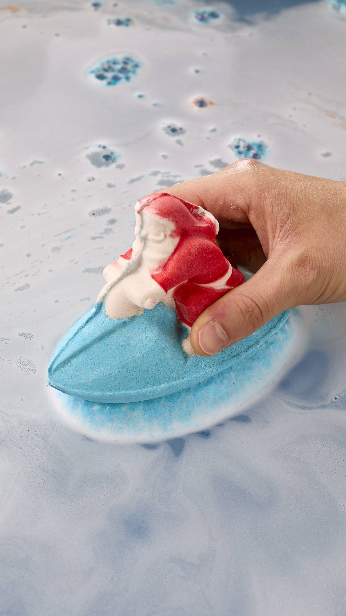 A hand holds a bath bomb shaped like Santa on a bright blue jet ski, placing it into pale blue bathwater where it releases a layer of white froth around its base.